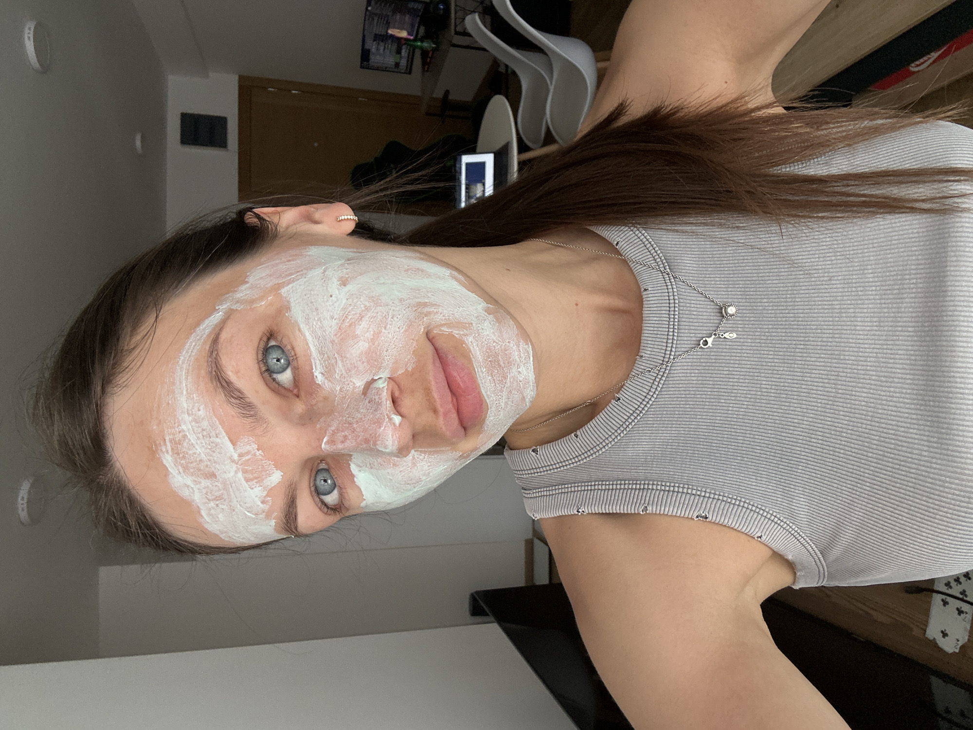 A woman is posing for a selfie indoors. She is wearing a grey striped tank top and a silver necklace with small charms. A white face mask is applied to her face. Her hair is brown, and her eyes are blue. In the background, there are white chairs and a computer. The location is indoors, presumed to be Donji Pobori, Montenegro.