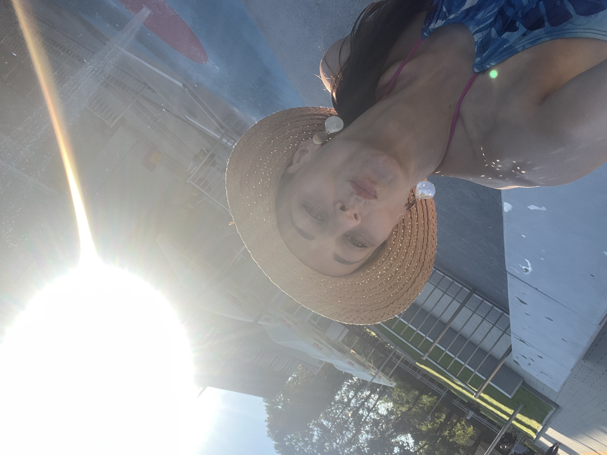 This is an outdoor, eye-level, medium shot of a person wearing a straw hat and a patterned bikini, with the image rotated 90 degrees clockwise. The person is looking towards the camera with a neutral expression. The sun is shining brightly, causing a significant lens flare that obscures the background. The person appears to be near a body of water, possibly a pool, given the water droplets and reflections visible on the surface. In the background, there are glimpses of buildings, trees, and some outdoor seating areas, suggesting a resort or a public leisure space. The time of day appears to be sunny, likely midday or afternoon, due to the strong sunlight. There are no discernible emotions beyond a neutral gaze. Some text is visible on awnings in the background, including "TYSKIE." The overall impression is of a person enjoying a sunny day outdoors.