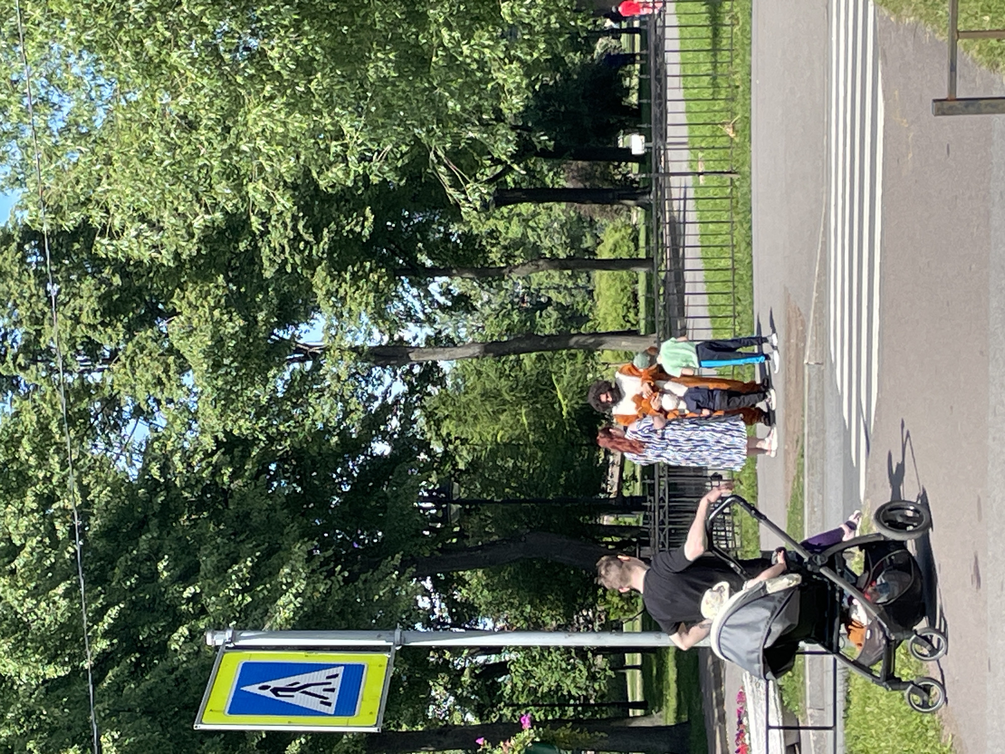 The image depicts a sunny day scene in a park with lush green trees and a pedestrian crossing. In the foreground, a man with short brown hair is pushing a stroller with a baby inside. The baby is wearing a light-colored bonnet. The man is wearing a black t-shirt and dark pants, with purple leggings visible beneath his pants. To his right, a woman with long red hair is wearing a patterned dress, and next to her, a child in a green t-shirt and black pants with blue stripes stands. Behind them, a person in a full-body costume, resembling a lion or fox, is holding what appears to be a stuffed toy or a puppet. A pedestrian crossing sign is visible on the left side of the image. In the background, a black metal fence separates the park area from a grassy field. The overall environment suggests a public park in the city of Saint Petersburg, Russia, likely during warmer months due to the abundance of foliage and clear skies.