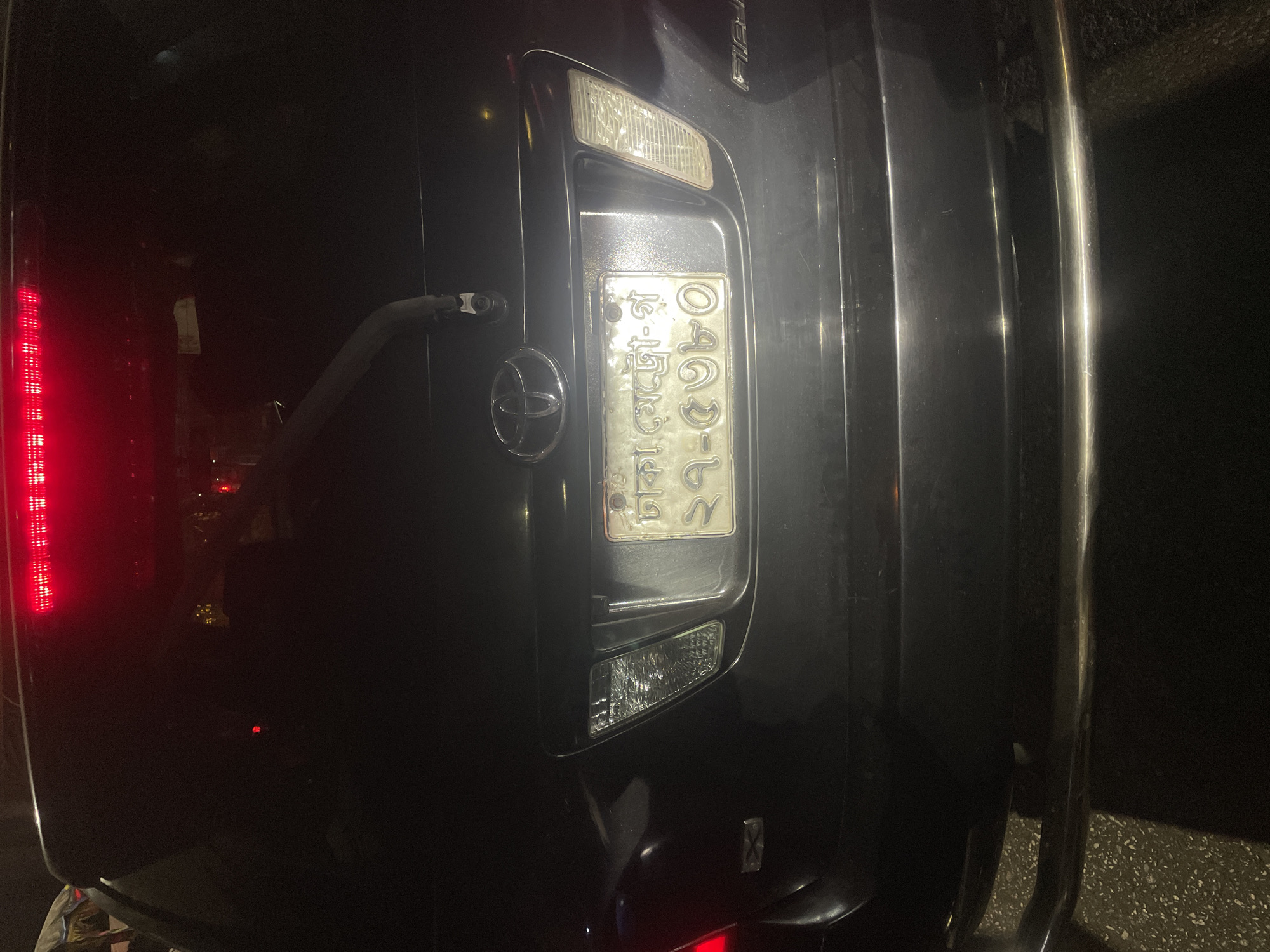 The image shows the rear of a dark-colored SUV, possibly a Toyota, at night. The vehicle's license plate is visible, bearing Bengali script, placing it most likely in Bangladesh, specifically likely Dhaka. The license plate appears to have the number "0903-5" visible on it. The left taillight is illuminated. The vehicle also has a metal bumper guard. The environment is dark, suggesting an outdoor location at night. The image is captured at an oblique angle, with the camera tilted.