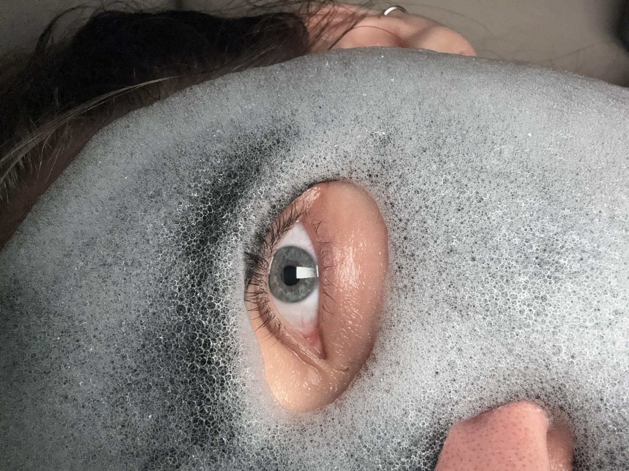 A close-up image captures a person partially submerged in a dense layer of white and grey foam, likely in a bath or spa setting. The central focus is a human eye and the upper part of an ear, emerging from the bubbly surface. The visible eye has a light blue-grey iris, a dark pupil, and long, dark eyelashes. A rectangular light reflection is visible in the pupil. The skin around the eye and the ear is pinkish, contrasting with the light foam.
Dark hair is visible at the top left of the frame, hinting at the person's head position. In the upper right, a hand or finger with a silver ring can be seen. The foam completely covers the rest of the person's face and body, creating a textured, undulating surface composed of countless small bubbles. The density and size of the bubbles vary, with some areas appearing more opaque white and others allowing a glimpse of darker areas beneath.
The person's eye appears calm and directly engaged, looking towards the viewer. The overall atmosphere conveyed is one of relaxation and tranquility. There is no visible text within the image. While the location context is given as Moscow, Russia, the image itself does not contain any specific visual cues to indicate this geographical detail. The scene suggests an indoor environment, with no explicit indicators of time of day or weather.