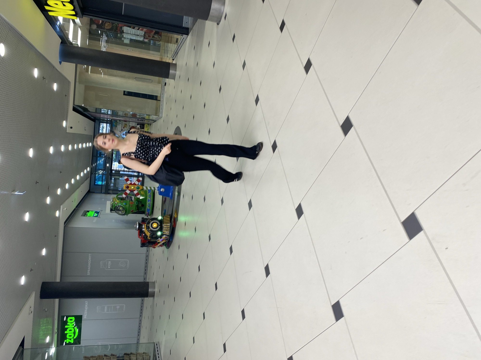 A young woman walks down a bright, tiled hallway in a shopping mall. She is wearing a black and white polka dot top and black flared pants, carrying a black bag over her left shoulder. In the background, a child's train ride is visible, along with store facades, including one with a yellow sign that says "New". The mall appears to be in Szczecin, Poland, suggested by the "Żabka" convenience store sign. The floor is tiled with large, light-colored squares, each with a small black square at its center.