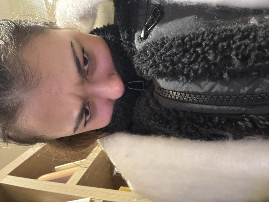 A woman looks into the camera with slightly furrowed brows. She wears a dark jacket with a zipper and fluffy black trim around the collar. There's white fur or stuffing around her shoulders. The photo is taken at an unusual angle, rotated so the woman appears sideways. Background features a wooden shelf unit with books or other items visible.