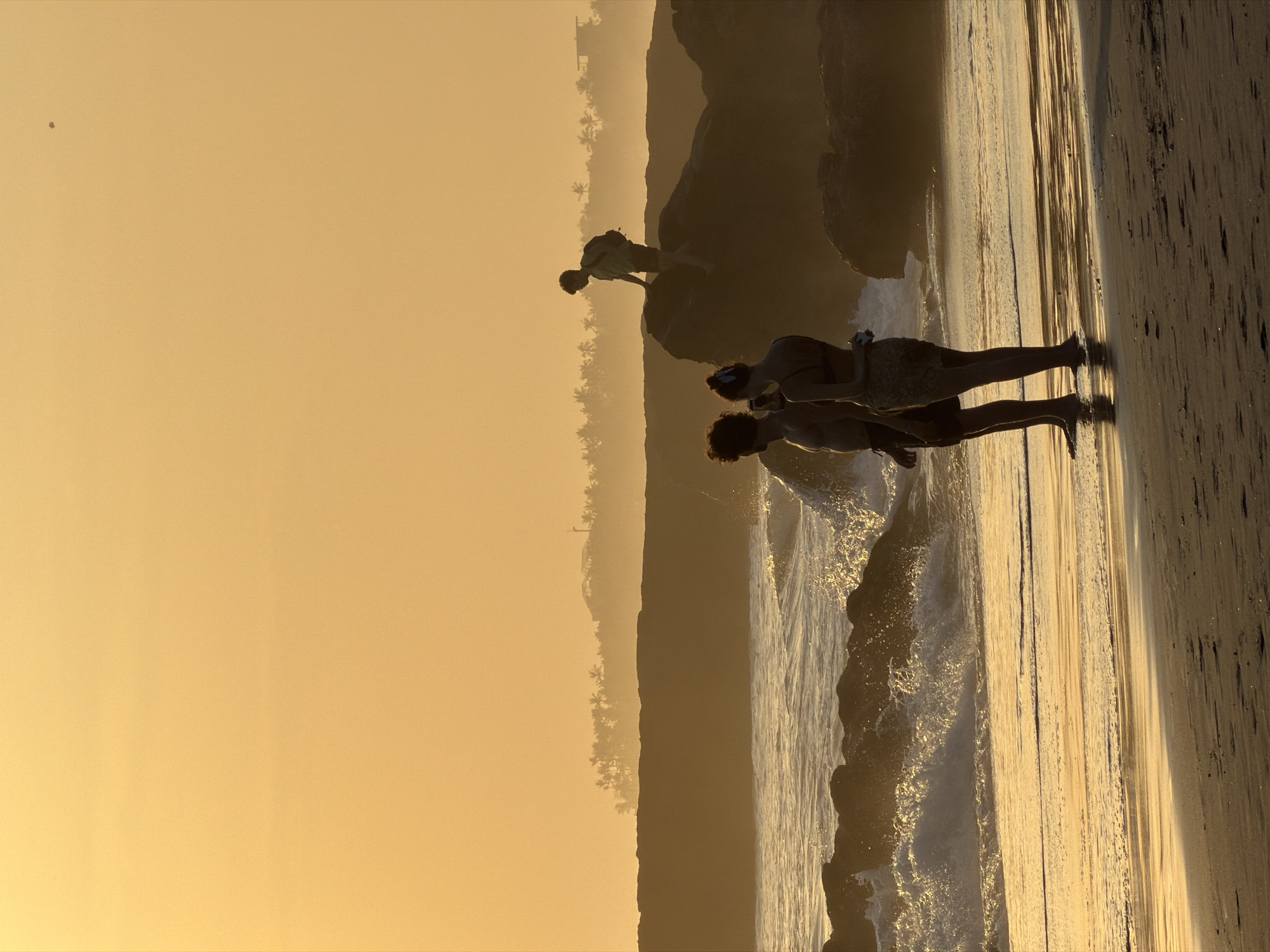 The image depicts a sunset scene on a beach in North Kuta, Indonesia. Silhouettes of three individuals are visible. In the foreground, a couple stands by the water's edge, with the male figure on the left and the female figure on the right, facing away from the camera. The female appears to be holding a phone. Further back, atop large rocks, a third individual, likely male with a backpack, is positioned looking out towards the horizon. The ocean is active with breaking waves, and a rocky shoreline is prominent. The sky is a warm, hazy orange, indicative of sunset, with a distant silhouette of palm trees and structures suggesting a coastal town. The beach itself shows wet sand reflecting the warm light.