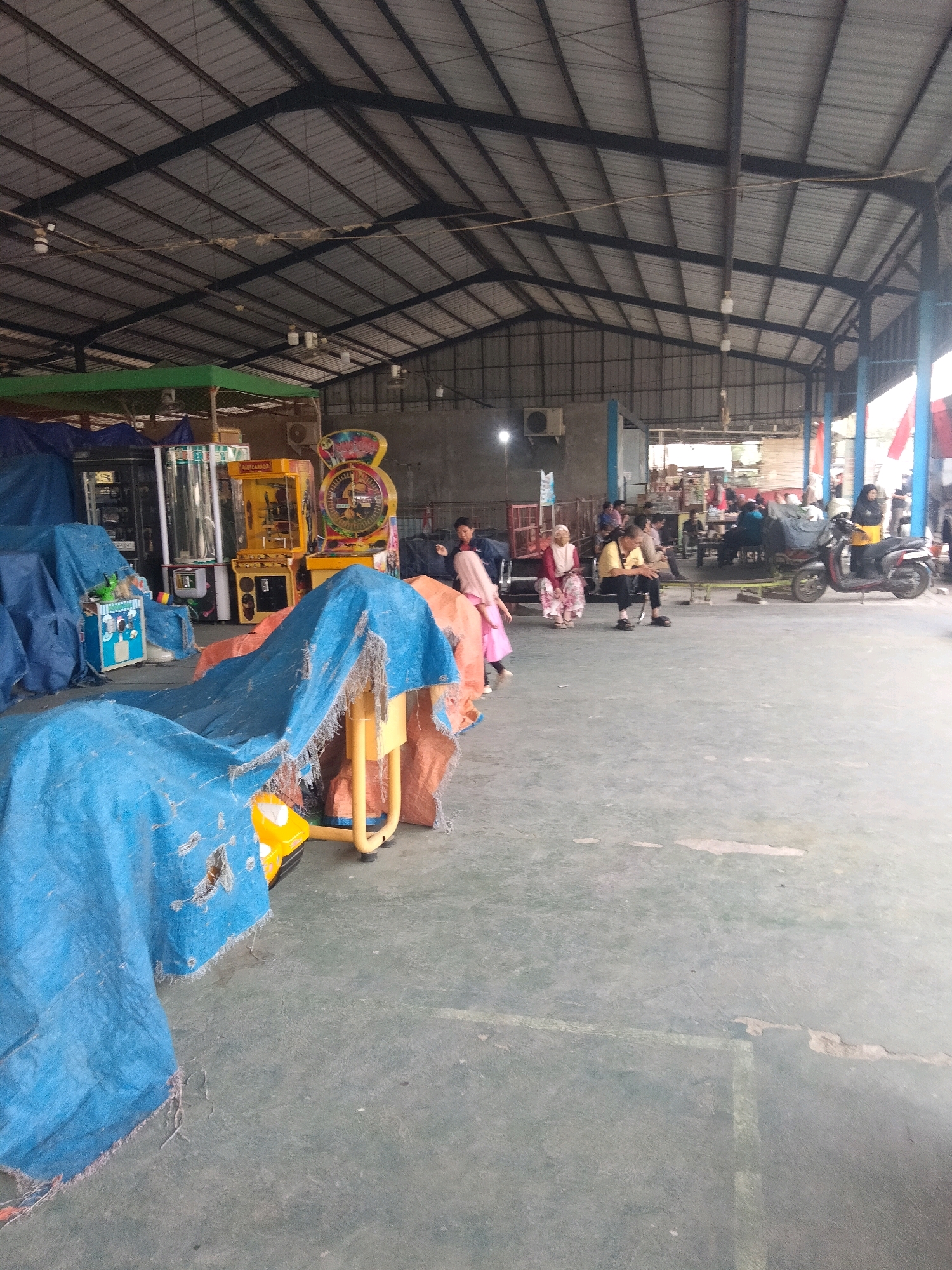 The image depicts an indoor area with a high, corrugated metal roof supported by dark metal beams. The floor is a gray concrete surface with some visible markings and minor damage. In the foreground, large blue tarpaulins cover various objects, one of which has a tear revealing an orange material underneath and a portion of a yellow toy.
To the left, several arcade machines are visible, including a prize claw machine with a clear cylinder, a yellow arcade game, and another game with a circular display. Some of these machines are also partially covered.
In the mid-ground, several individuals are seated or standing. Three women are seated on a bench to the left, one wearing a light pink headscarf and dress, another in a white headscarf and pink patterned attire, and a third in a light pink outfit. Further to the right, a man in a yellow shirt and black pants is seated on a bench. Behind them, more people are visible in the distance, some seated around tables. A red and white striped flag is visible on the right side of the frame.
A red scooter is parked on the right side of the image, with a person in a yellow shirt standing next to it.
The overall environment appears to be an open, covered space, possibly a marketplace or an outdoor recreational area with arcade attractions in Sukaasih, Indonesia. The presence of arcade machines and seating areas suggests a place of entertainment.