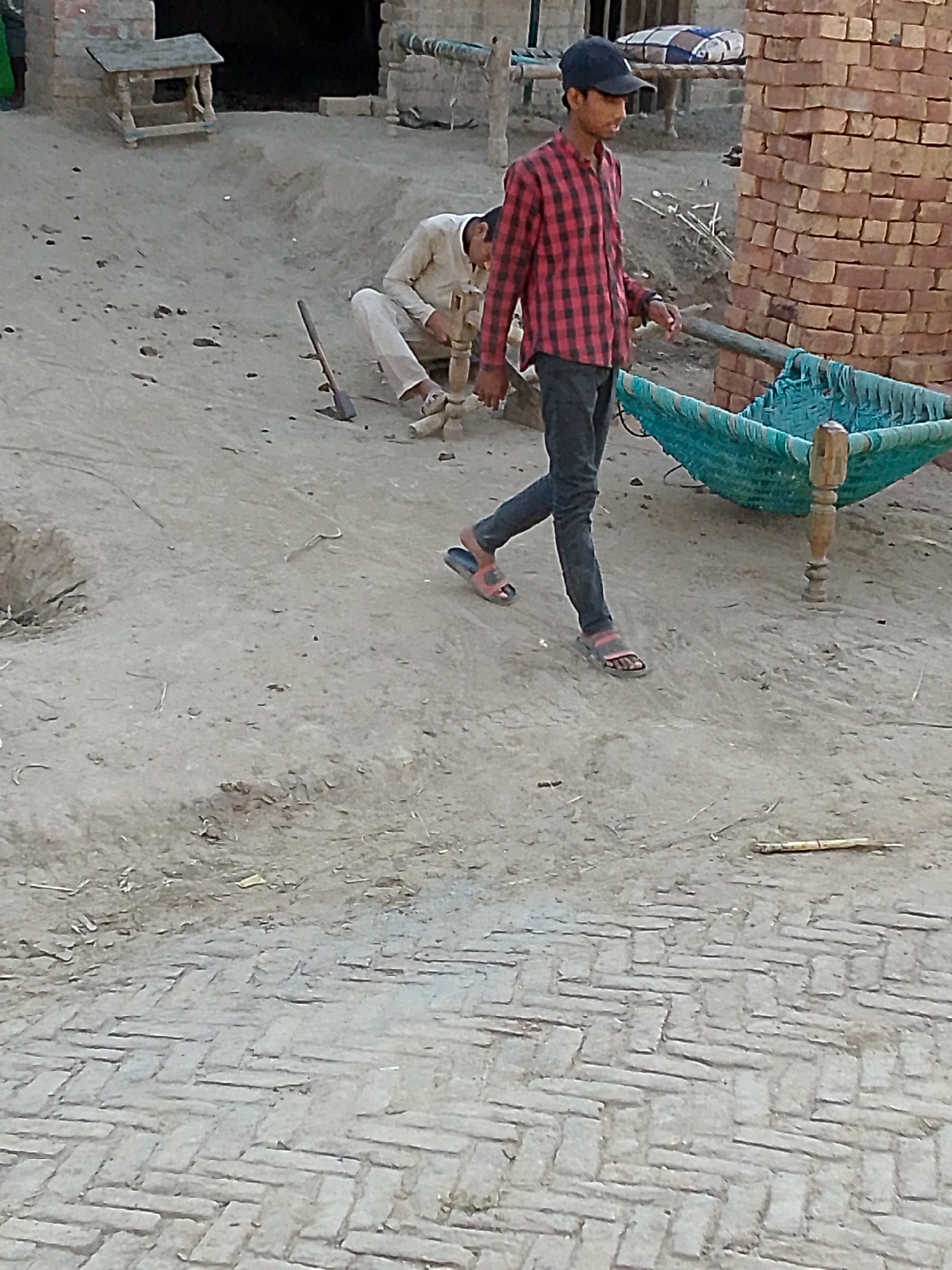 The image depicts an outdoor scene in what appears to be a rural or village setting. In the foreground, a young person wearing a red and black plaid shirt, dark jeans, a dark baseball cap, and sandals is walking. To their right is a traditional charpai bed with blue woven netting. Behind them, another person is kneeling, possibly working on something. An ax is lying on the ground nearby. The background includes structures made of brick and mud, with various objects scattered around, including a small wooden table-like structure and another bed. The ground is dirt and brick. The location is in a city of Unknown, Unknown.