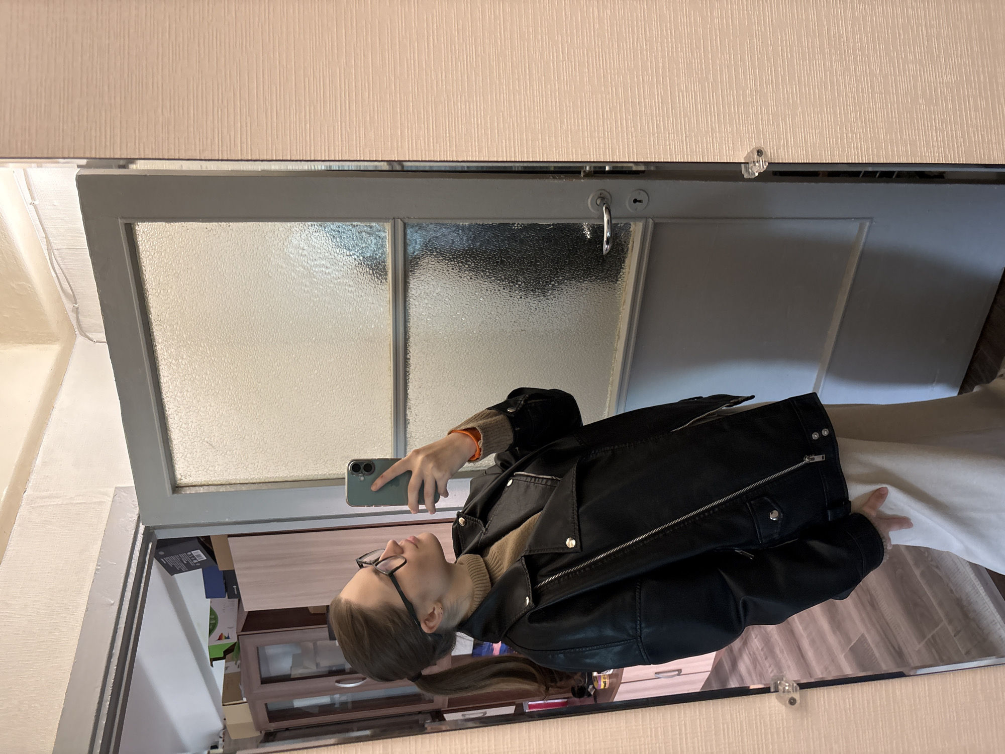 A woman is taking a selfie in a mirror in a room. The room appears to be in a residential setting. The mirror is mounted on a door, possibly a closet door, and has frosted glass panes in the upper section. The woman is wearing glasses, a tan turtleneck sweater, and a black leather jacket. She is holding a smartphone in her left hand to take the photo. The mirror reflects the inside of the room, including a cabinet and various objects on shelves. The walls are light-colored. The location is Saint Petersburg, Russia.