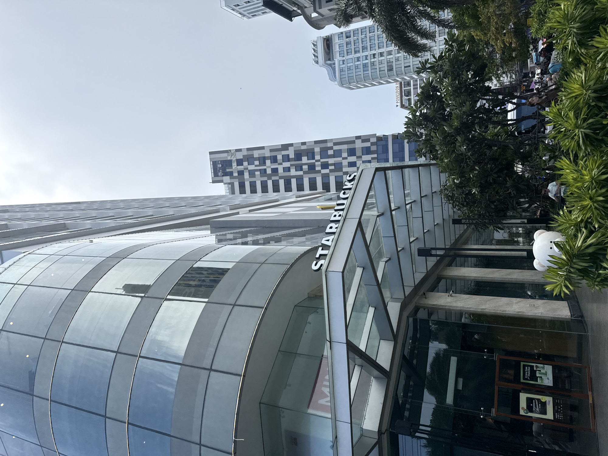 The image captures a modern urban scene in what appears to be Nha Trang Ward, Vietnam, focusing on a Starbucks coffee shop. The Starbucks is housed in a contemporary building with a prominent curved, glass-paneled facade on the left and an angular glass entrance on the right. The "STARBUCKS" sign is vertically displayed along the angular glass section.
In the background, several tall, modern buildings stand against a bright, clear sky. One building directly behind the Starbucks features a distinctive checkerboard pattern of light and dark panels. To the upper right, another building has "POSEIDON H" visibly inscribed on its side, suggesting "Poseidon Hotel."
Lush green trees and foliage are abundant, framing the right side of the Starbucks entrance and appearing in the mid-ground, where faint figures of people can be seen. A large, white, bear-shaped head sculpture or balloon is placed near the Starbucks entrance, partially obscured by plants. The Starbucks entrance comprises large glass doors with visible signs, including a "STARBUCKS REWARDS" logo, promotional material for beverages (featuring green drinks), and text indicating opening hours such as "Mondays-Fridays." A partial reflection or sign showing "MIN" can be seen on the curved glass of the building. The scene depicts daytime with an evenly lit sky, suggesting either an overcast day or early morning/late afternoon.