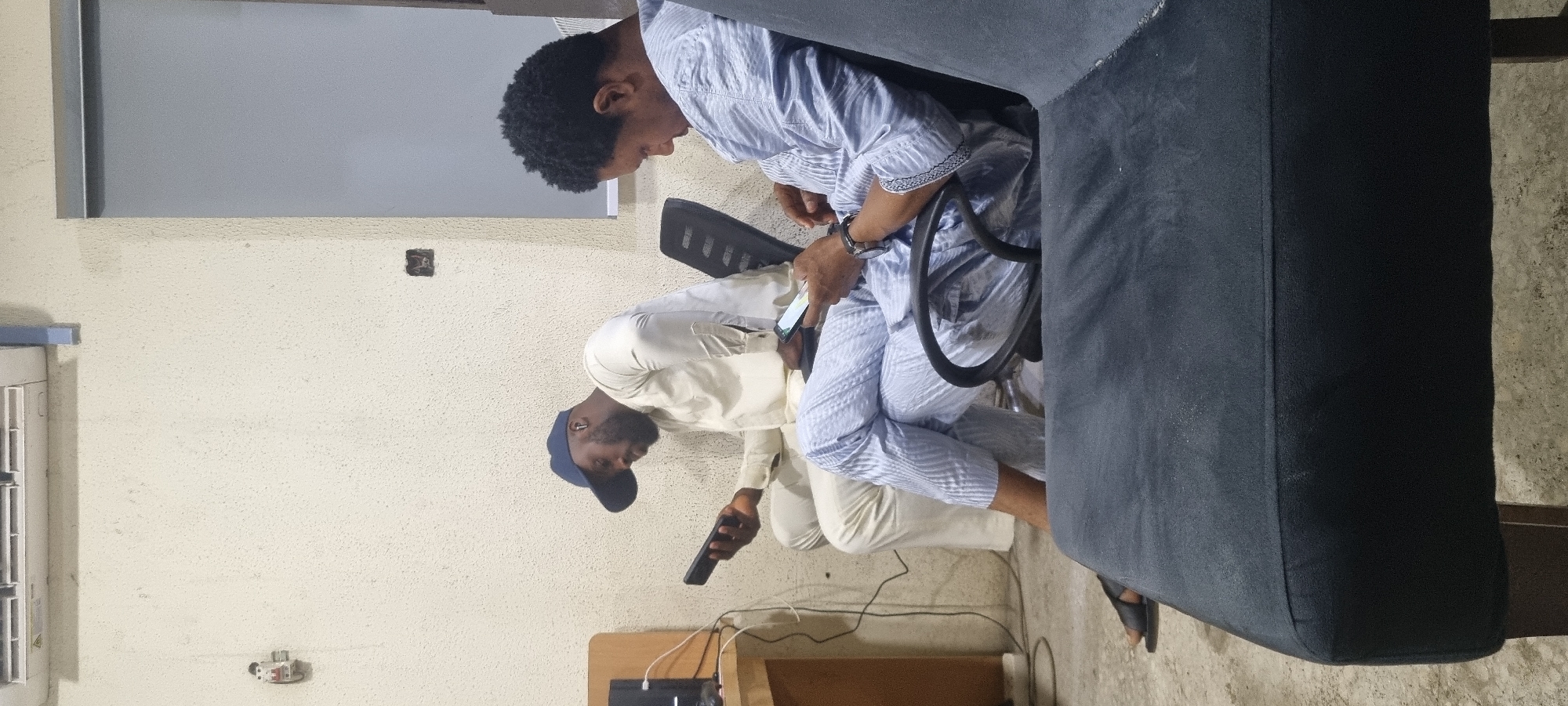 The media file depicts two individuals seated in what appears to be an indoor setting, possibly an office or waiting area. The scene is oriented sideways, with the subjects appearing to be upright.
The first individual, on the left, is wearing a light-colored traditional outfit and a dark blue baseball cap. They are holding a dark rectangular object, possibly a remote control or a phone, in their right hand and looking down at it. They are seated in front of a beige, textured wall.
The second individual, on the right, is wearing a light blue and white striped patterned outfit. They are seated in a black office chair and are also looking down at a mobile phone they are holding in their right hand. A dark gray couch or seating area is visible to the right of this individual.
The background shows a light-colored textured wall with some electrical outlets and a conduit. A window with a light gray blind is partially visible at the top of the frame. Cables are seen on the floor, connecting to what might be electronic equipment. The overall lighting suggests it is daytime, but the indoor setting makes it difficult to determine the precise time of day or weather conditions.
The overall mood appears relaxed, with both individuals engaged with their electronic devices. No visible text is present in the image. The context provided places the location as Ikeja, Nigeria.