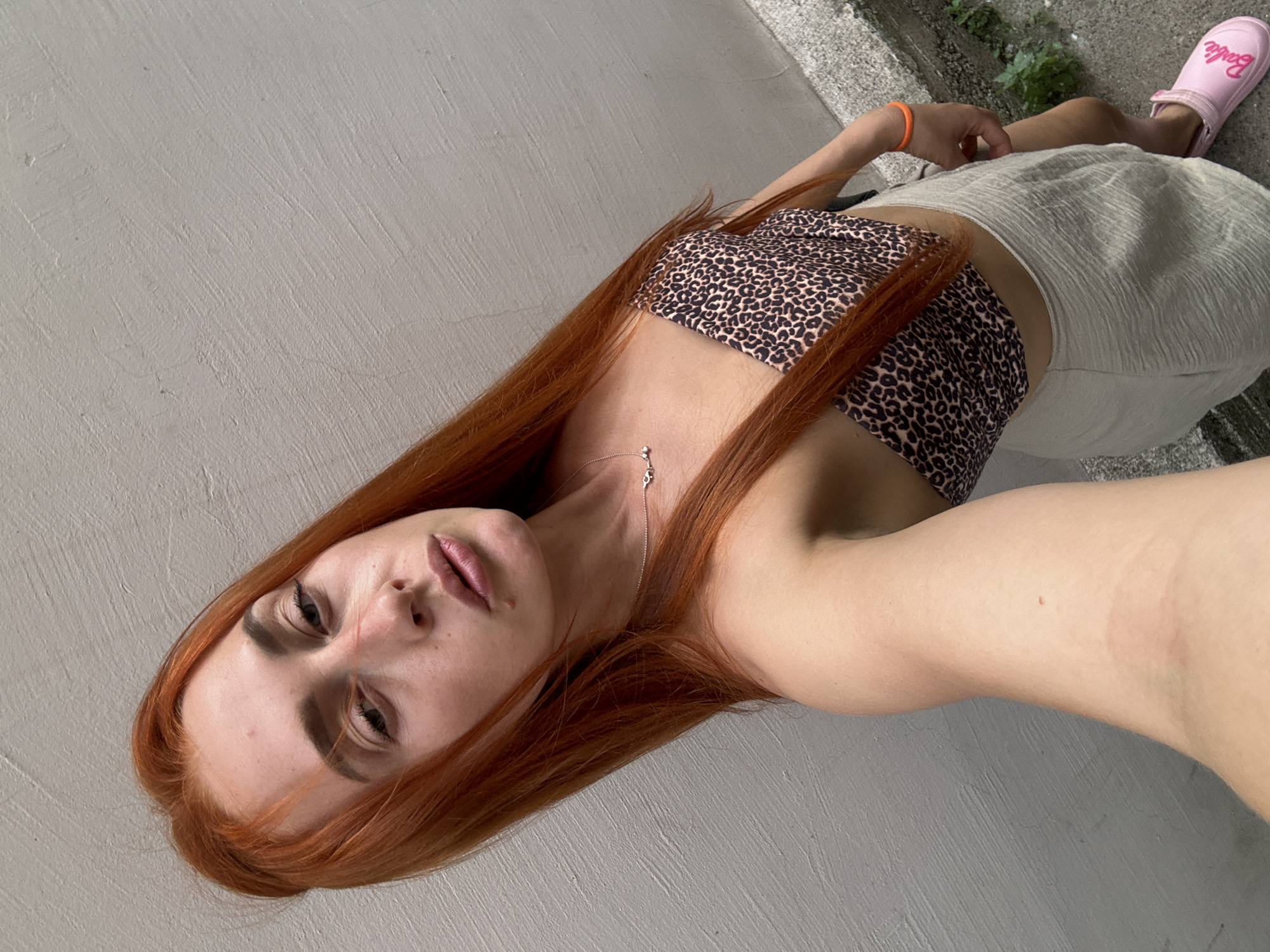 A young Caucasian woman with long, bright red hair is standing in front of a textured, off-white wall, possibly outdoors in Budva, Montenegro, as indicated by the context. She is wearing a leopard-print bandeau top and a light-colored, textured skirt. Her left hand is holding the camera, as she is taking a selfie. On her right wrist, she is wearing a bright orange bracelet, and her right foot is visible wearing a pink "Barbie" branded clog. The background wall appears to be slightly weathered, and to the right, a concrete surface with some scattered greenery is visible. The lighting suggests it is daytime.