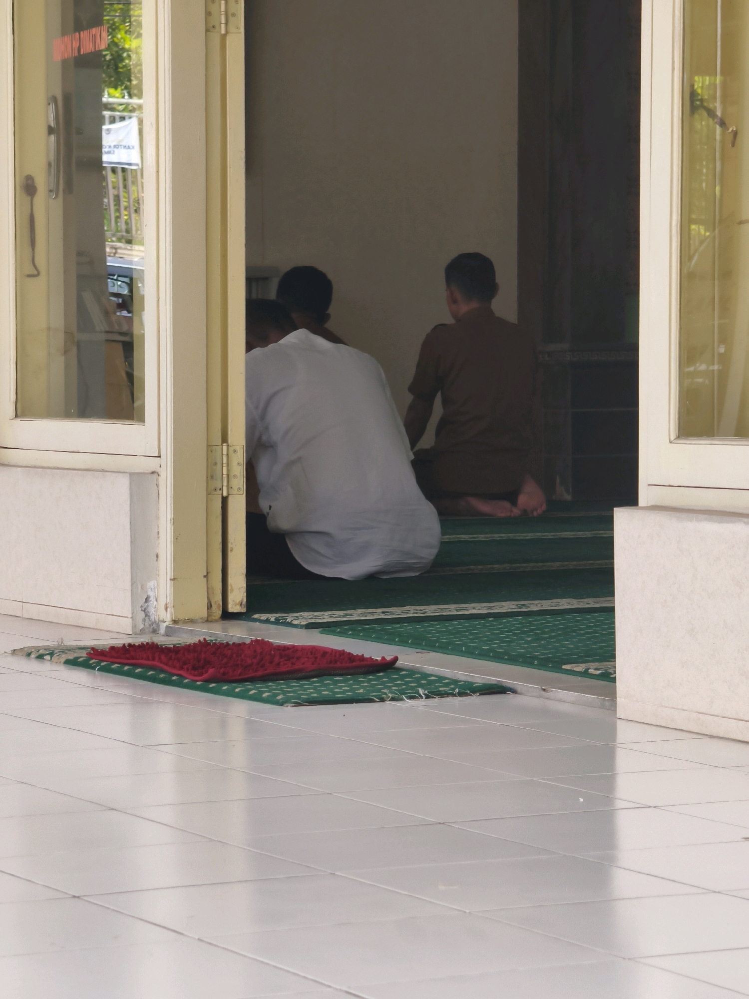 The image shows two men praying on prayer mats inside a building, likely a mosque or prayer room, located in Samarinda, Indonesia. The building appears to have a white tiled floor in the foreground and a green carpeted area where the men are praying. One man is wearing a white garment, and the other is wearing a brown shirt. They are both in a kneeling position, facing away from the camera. Through an open doorway, a glimpse of the outside world can be seen, including a white fence and trees. The doorway is framed by a yellow structure, and a glass-paneled door is visible on the left. A small red mat is placed on the white tiled floor in front of the entrance to the prayer area. The lighting suggests it is daytime.