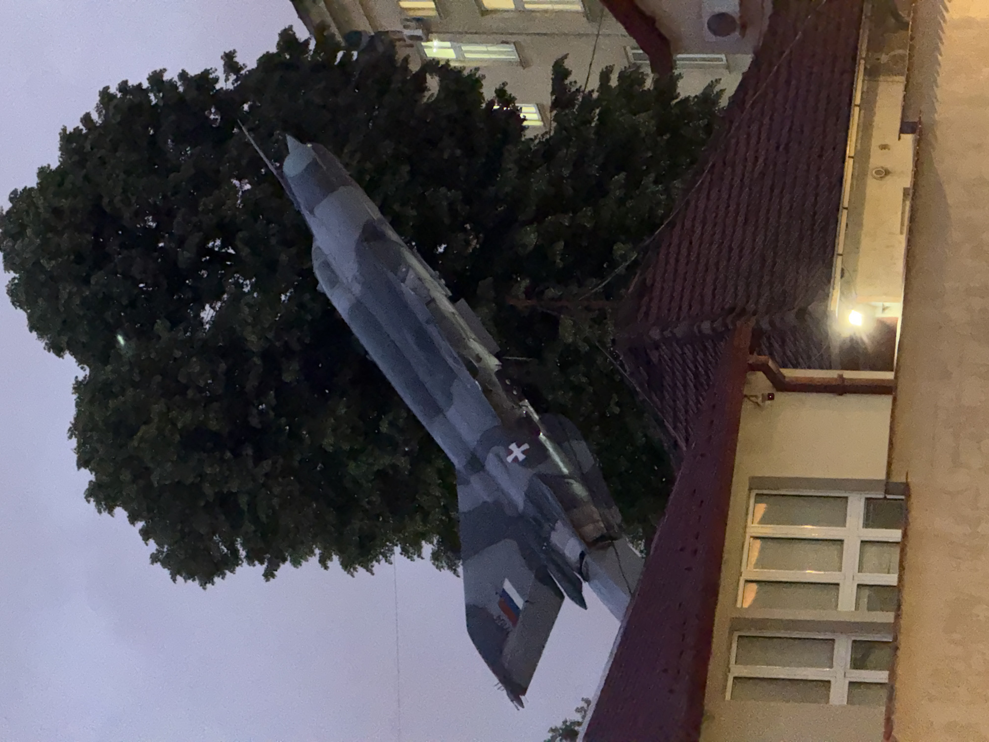 An image shows a Mikoyan-Gurevich MiG-21 aircraft, mounted as a static display, in Belgrade, Serbia. The aircraft is gray camouflage and bears the red star emblem of the Yugoslav Air Force and the Serbian flag. It is positioned adjacent to a building and beneath a large tree. The building has multiple white-framed windows. The image appears to be taken during dusk or evening, as evidenced by the dim lighting.