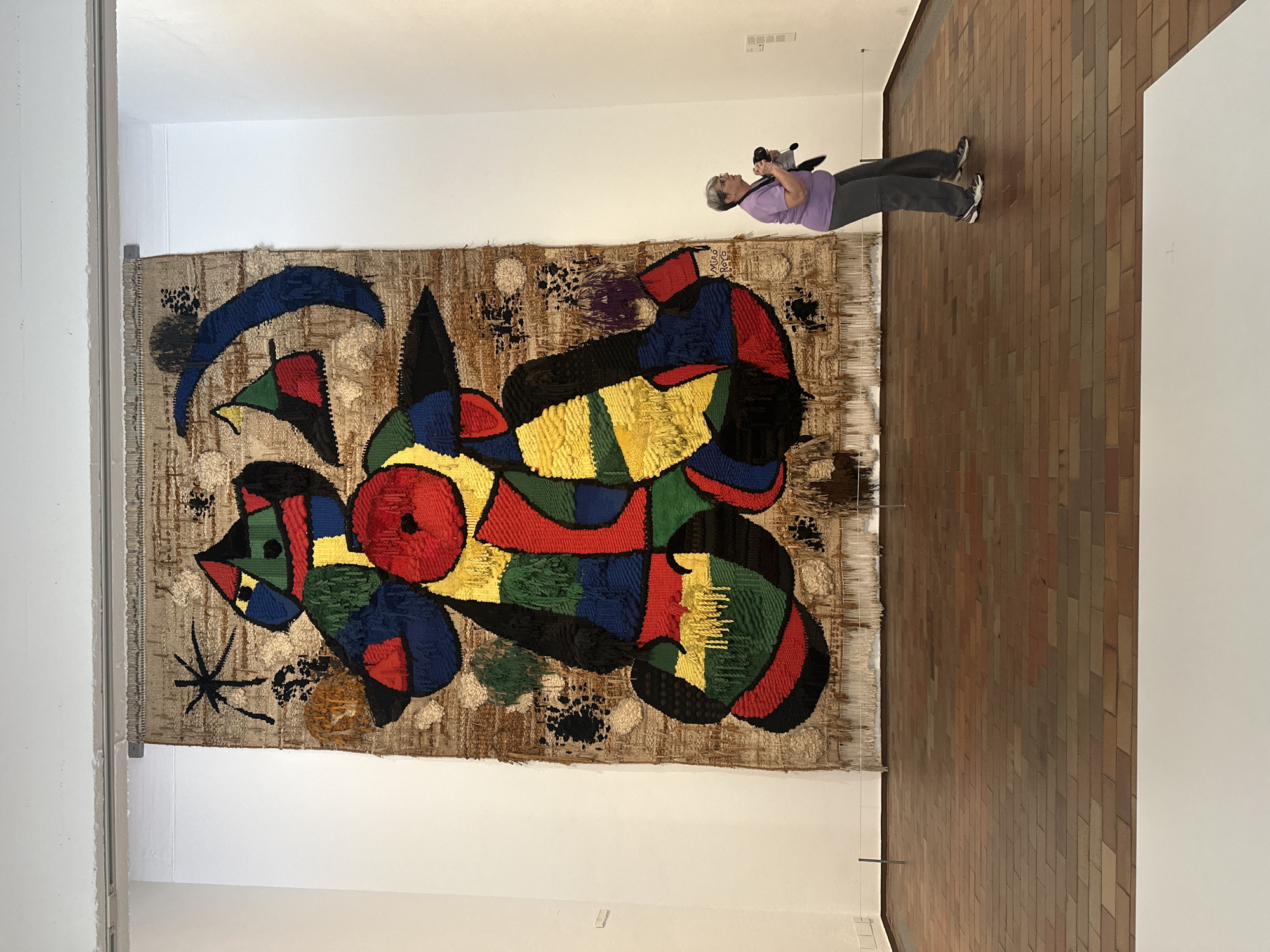 A horizontal, eye-level shot captures a museum setting in Barcelona, Spain. The main focus is a large, colorful tapestry, an artwork by Joan Miró named, "Barcelona Tapestry". The tapestry displays abstract, geometric shapes in red, yellow, blue, green, and black on a beige background. It's hanging against a white wall.
To the right of the tapestry, a woman is seen holding a camera and seemingly taking a picture of the artwork. She has short, gray hair, a purple t-shirt, and gray pants. The woman is positioned in front of a wall covered in brown, rectangular tiles. The tiles are arranged in a brick-like pattern.
The environment is well-lit, typical of a museum setting. The walls are painted white and a track lighting system can be seen on the ceiling. There's also a metal railing on the side of the tapestry.