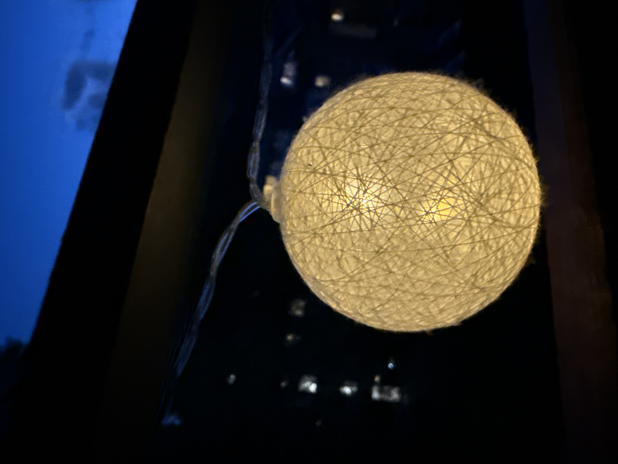 The image depicts a close-up, angled shot of a spherical string light, illuminated from within by a warm yellow glow. The string light is made of interwoven off-white threads, creating a textured, spherical surface. A thin, transparent wire extends from the top of the sphere.
The background is dark, with blurred out lights suggesting an urban or suburban setting at night. The upper portion of the image shows a deep blue sky with faint clouds. The overall impression is one of a cozy, possibly outdoor setting at dusk or night.
While the image itself does not provide specific visual cues to pinpoint the exact location within Garmisch-Partenkirchen, Germany, the presence of city lights in the background and the twilight sky are consistent with an urban environment. The string light is likely a decorative element on a balcony, patio, or outdoor seating area. There are no people or identifiable landmarks present in the image.