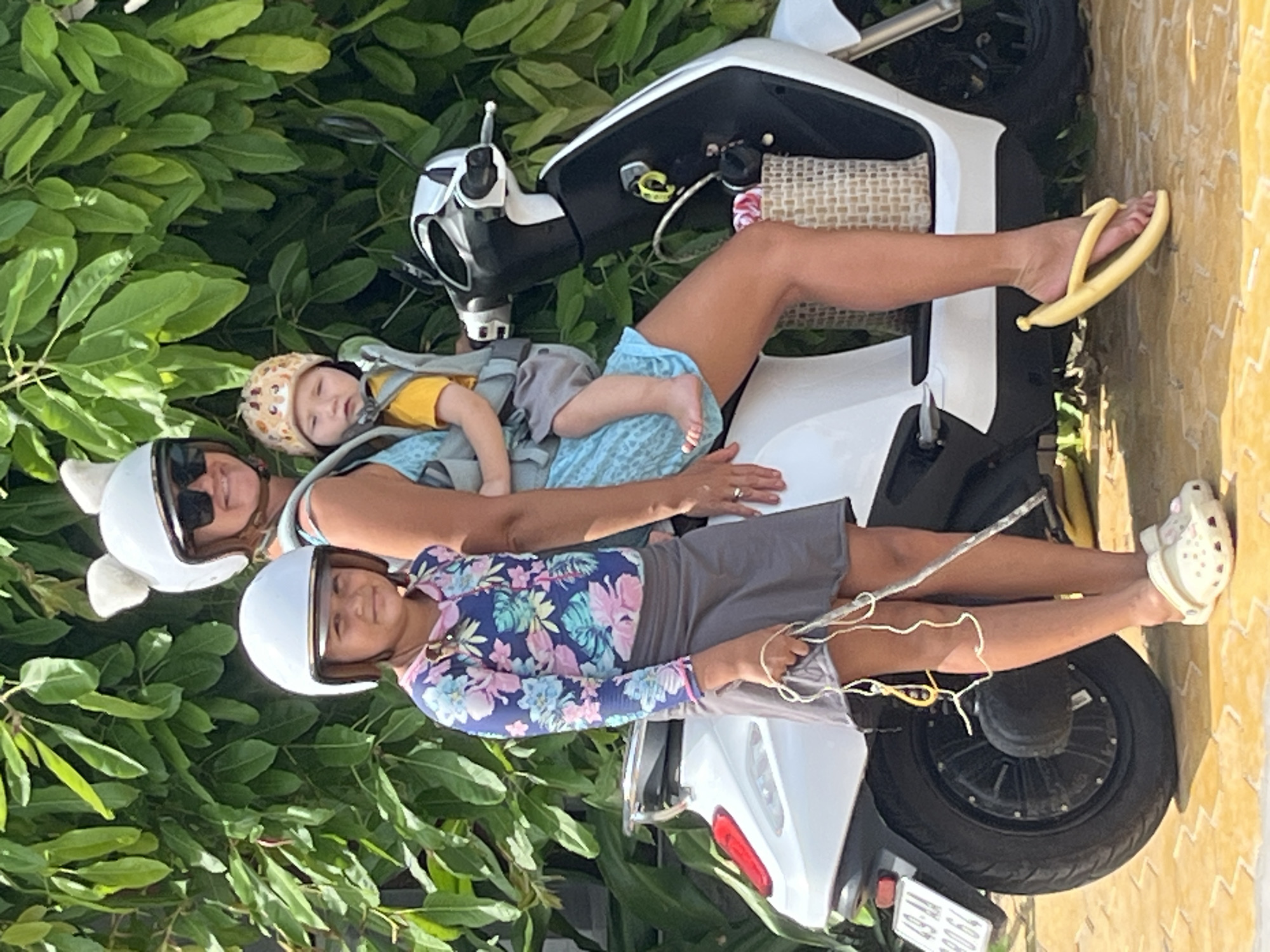 In Mũi Né, Vietnam, a woman wearing a white helmet with fluffy cat ears and sunglasses, stands next to a white scooter. She is holding a baby in a gray carrier attached to her front. The baby wears a patterned hat and a yellow shirt. To the left of the woman and slightly in front of the scooter, a young girl, wearing a white helmet and a long-sleeved floral rash guard and a grey skirt, stands holding a jump rope. The scooter has a license plate that reads "49-AA 090.64". Behind the subjects is a dense green foliage. The ground is paved with light-colored square tiles. The woman is wearing yellow banana-shaped flip-flops, and the girl is wearing white clogs.