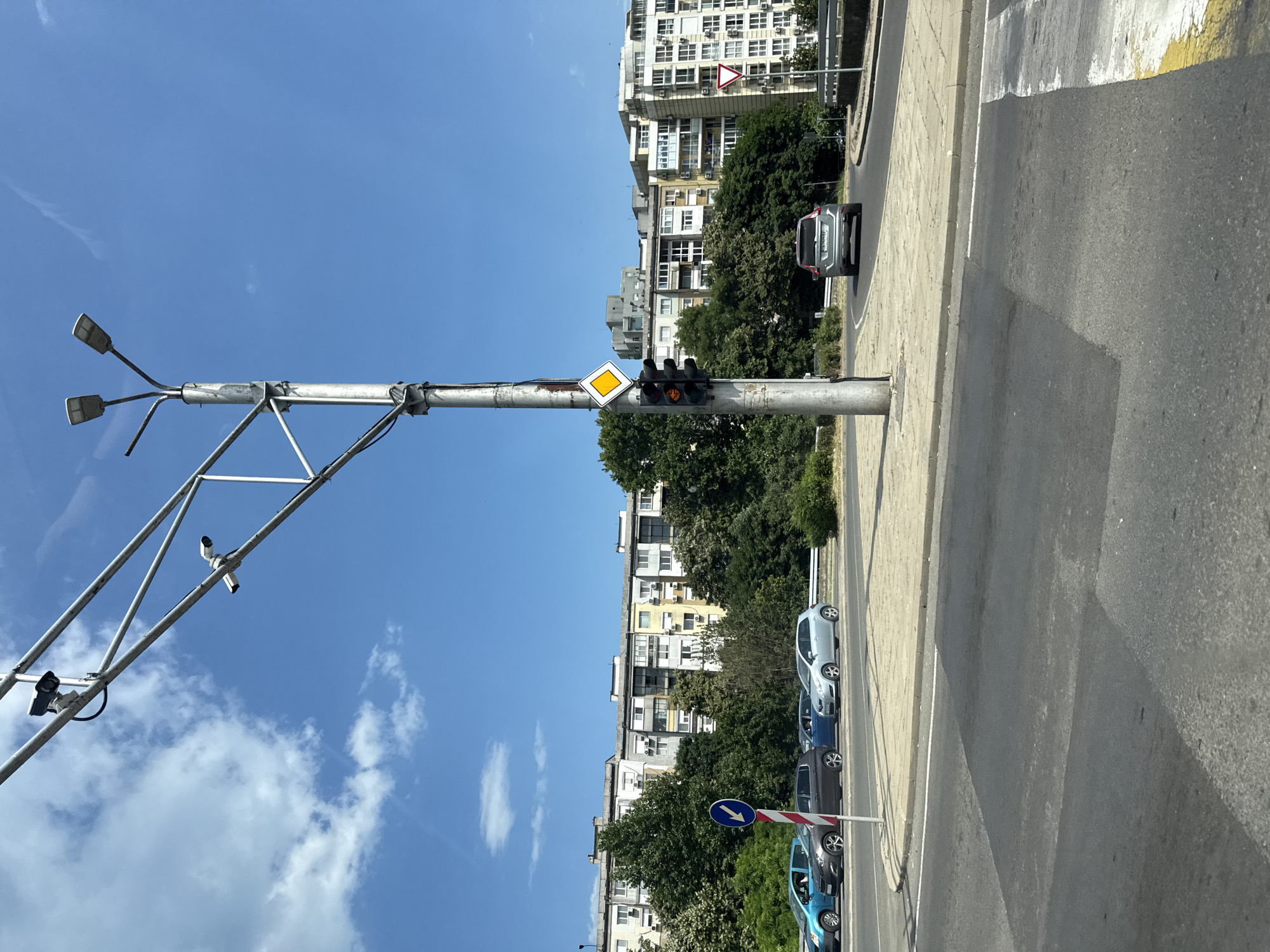 This is a medium shot, eye-level photograph of an urban street scene in Burgas, Bulgaria, under a clear blue sky with scattered white clouds. The primary focus is a utility pole with traffic lights, streetlights, and two security cameras mounted on a metal structure above. The traffic light is illuminated with amber. A diamond-shaped yellow sign is positioned above the traffic signal. The pole is situated on a sidewalk adjacent to a road.
In the background, a row of apartment buildings with balconies and visible windows stretches across the frame. Trees and greenery are interspersed between the buildings and along the roadside. Several cars are visible on the road, including a blue hatchback on the left, a dark grey hatchback, a light blue sedan, and a silver SUV in the distance turning to the right. A pedestrian crossing is visible in the foreground. A circular blue sign with a white arrow pointing left and a red and white striped pole are also present on the sidewalk. A triangular white sign with a red border and a red cross shape is visible on the right side of the frame, near the road. The overall environment suggests a typical city street during daytime.