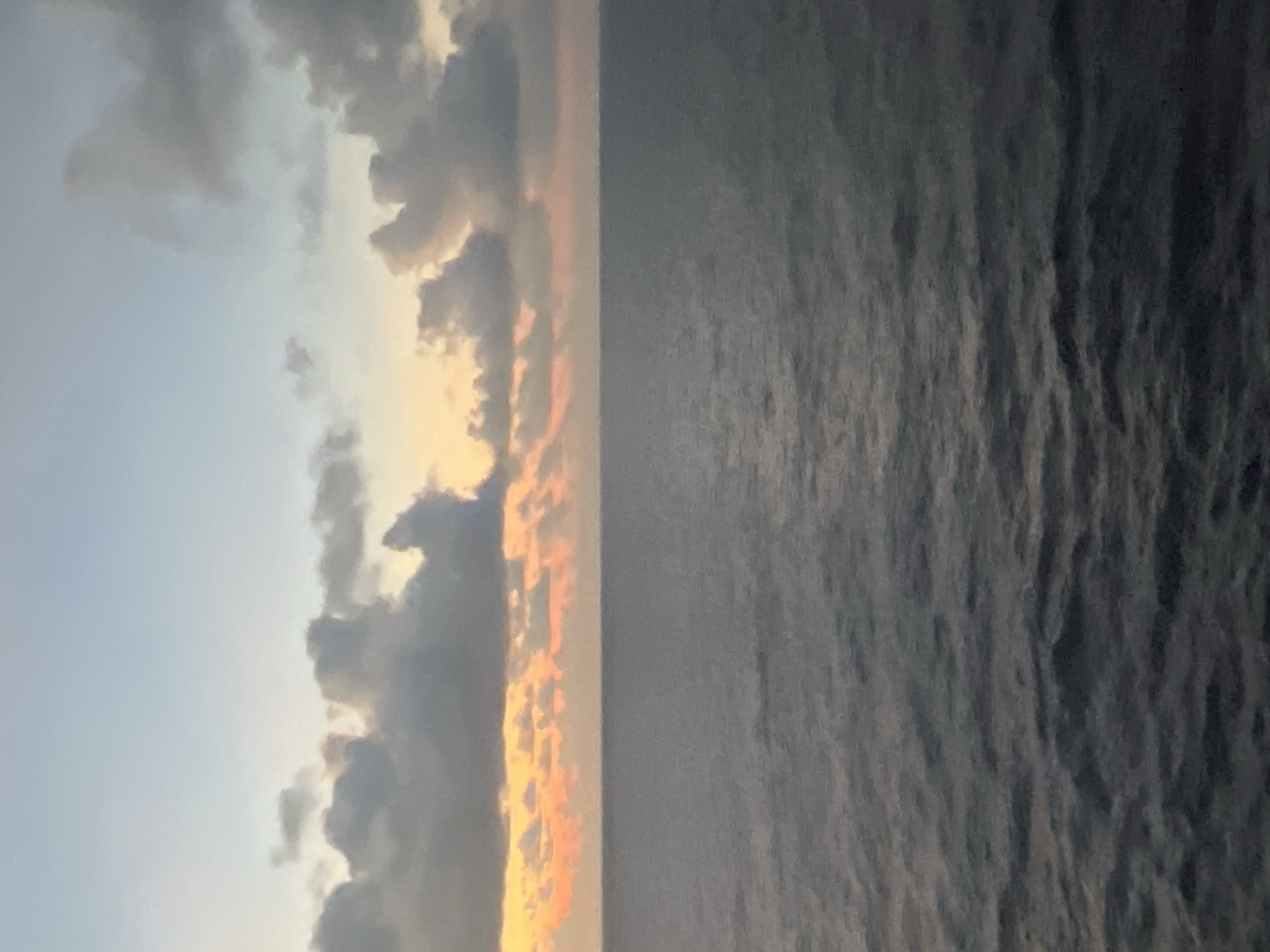 A wide shot of the ocean at dawn captures the transition from night to day with a mix of dark, choppy water and a sky painted with soft hues of orange and grey. The horizon line, where the water meets the sky, is a thin, indistinct band. In the upper portion of the image, scattered clouds drift across a pale blue sky, tinged with the warm glow of the rising sun. The water itself is a deep, muted blue-grey, with subtle ripples suggesting gentle waves. The scene is devoid of any human presence or man-made structures, creating a sense of vastness and solitude.