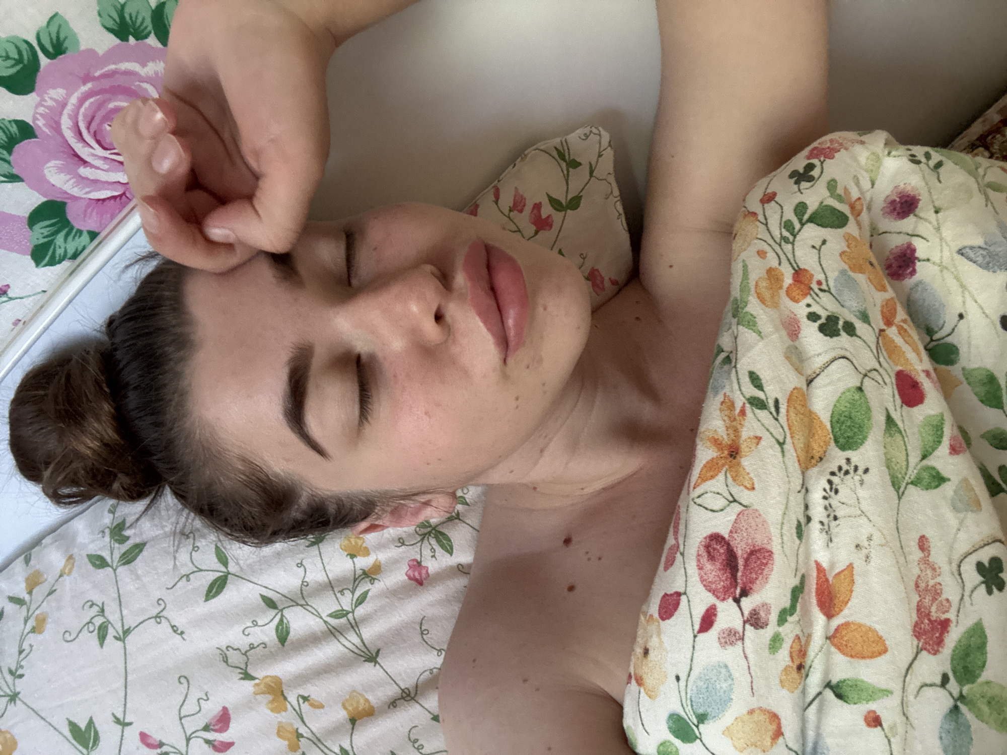 A woman is lying in bed. She is on her back with her eyes closed and a hand resting on her forehead. Her hair is pulled back in a bun. The bed is covered in a floral-patterned sheet and a floral-patterned pillow. There is no evidence in the image to suggest this is the city of Мещёры, Russia.