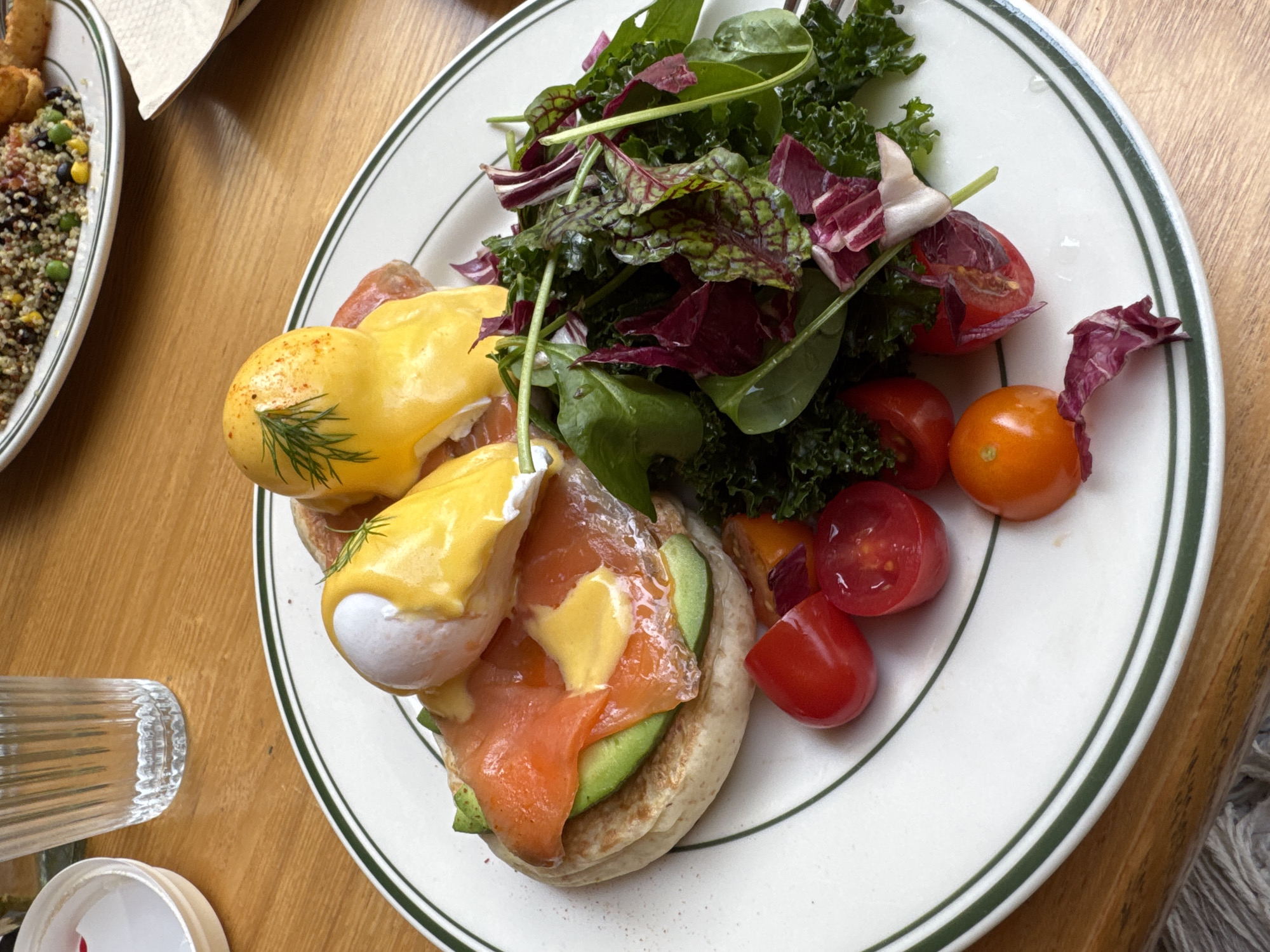 A plate of Eggs Benedict with salad sits on a wooden table. The plate has a white surface and is edged with a double green stripe. The Eggs Benedict consist of two poached eggs topped with hollandaise sauce, served on English muffins with avocado and smoked salmon. The salad includes kale, spinach, radicchio, and halved cherry tomatoes in red and orange varieties. A glass with some clear liquid is visible to the left of the plate. Part of another plate with a quinoa salad and a napkin is visible on the top left. The scene is well-lit, suggesting daytime. The context of being in Yuzhong District, China is unsupported by visual evidence.