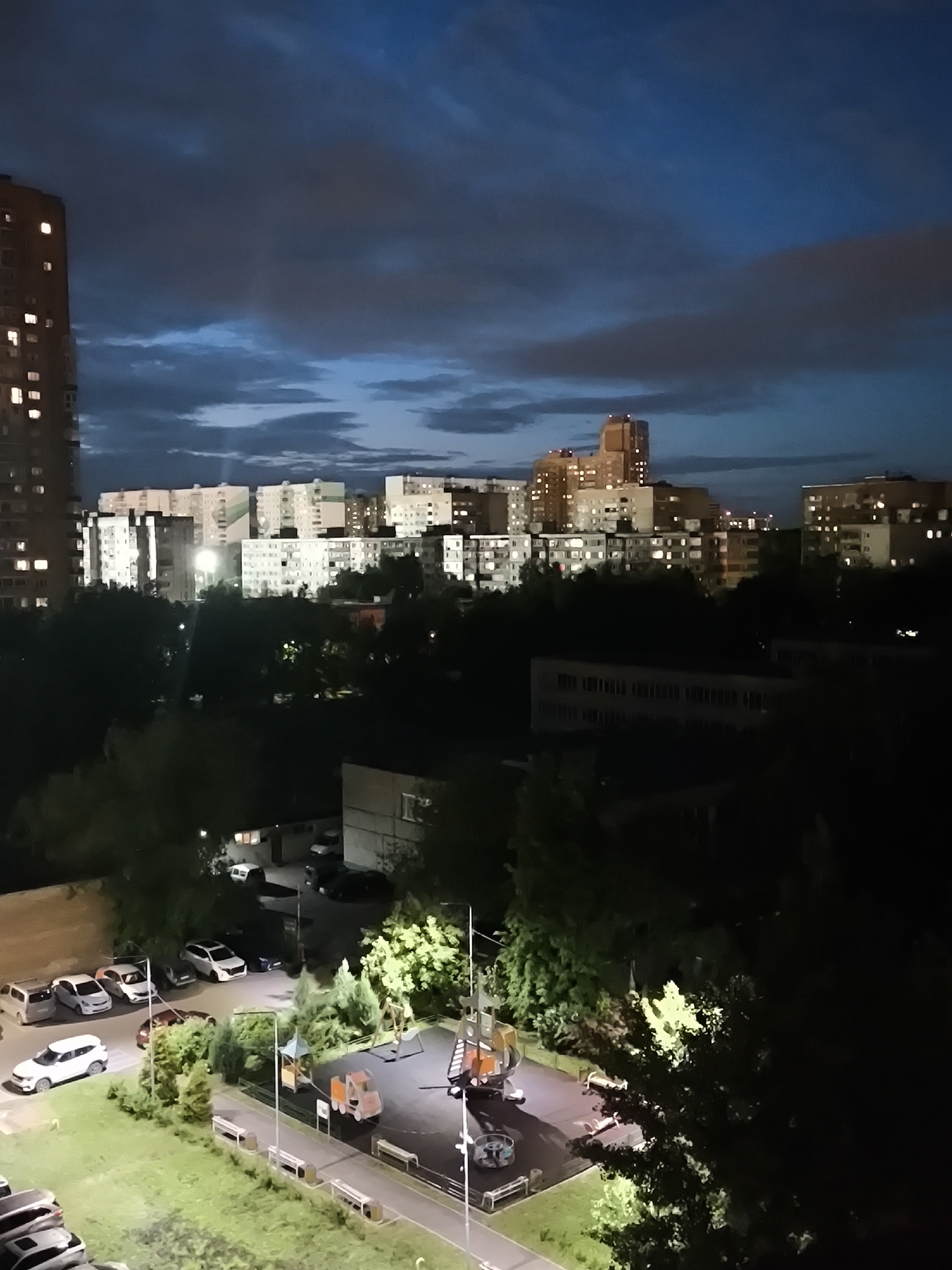 The image captures an urban landscape at night. In the foreground, there is a playground illuminated by a street light. Adjacent to the playground, a parking lot contains several parked cars. Beyond the trees and low buildings, there are taller apartment buildings, some with lit windows. The sky above is cloudy, with varying shades of blue.