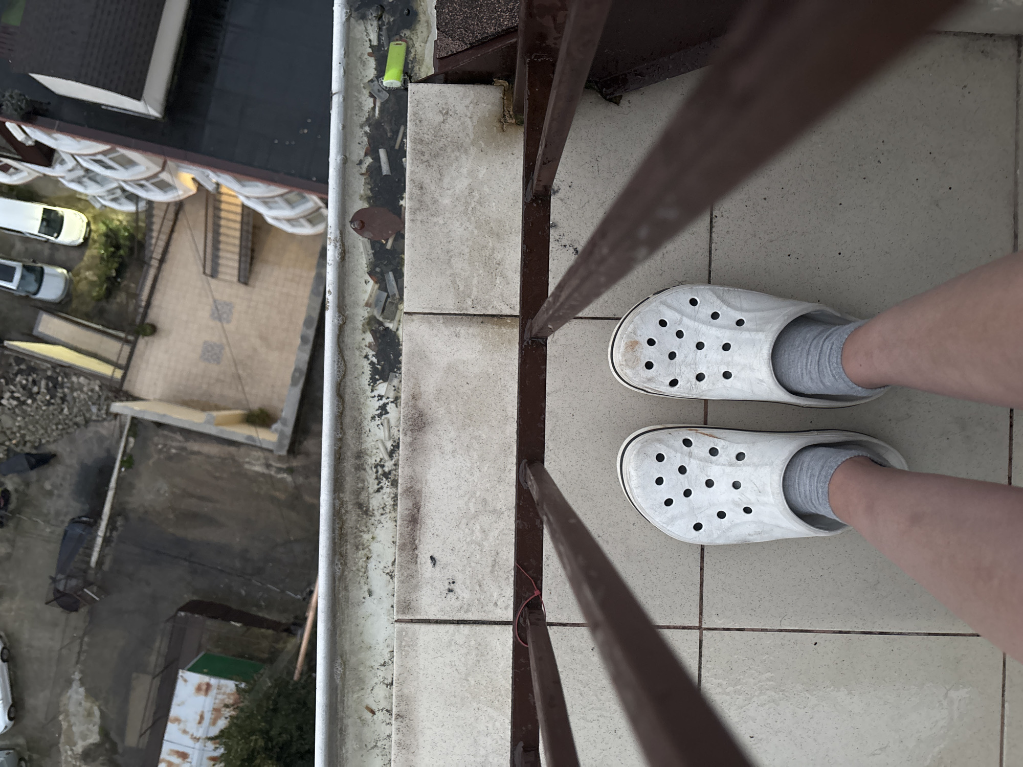 The image depicts a view from what appears to be a balcony overlooking a cityscape. The visible portion of the balcony features tiled flooring and a metal railing. A pair of feet wearing white Crocs and grey socks are positioned on the tiles, suggesting a person is standing on the balcony. The cityscape includes buildings with cars parked below. Debris and potentially construction material sit along the balcony's edge. The location is identified as Sochi, Russia.
