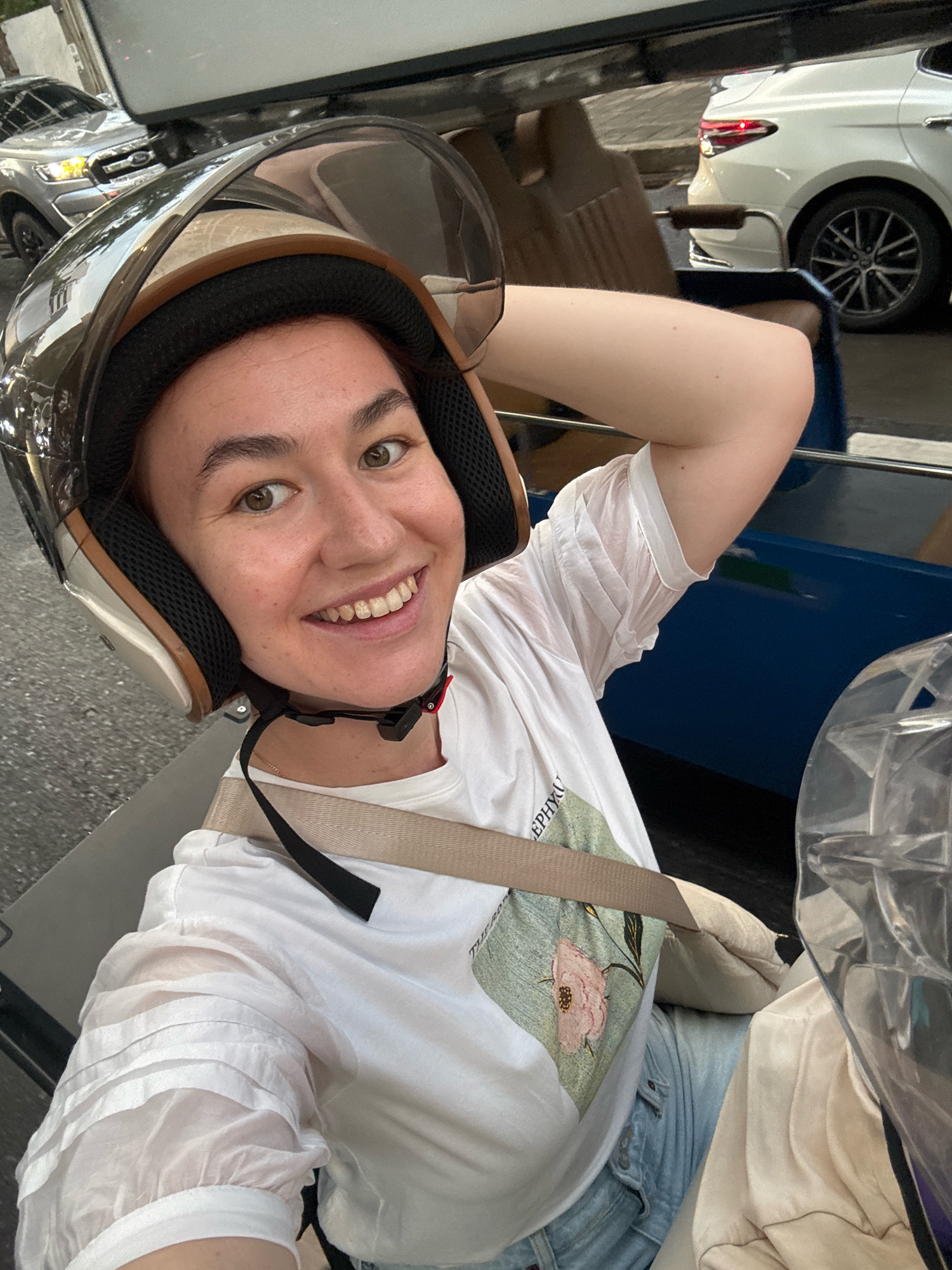 A young woman wearing a helmet and white t-shirt with a floral design is taking a selfie while riding in a tuk-tuk, which is a motorized rickshaw commonly used as a taxi in Bangkok, Thailand. The tuk-tuk is blue, and the woman is smiling at the camera. In the background, other vehicles, including a white car and a silver pickup truck, are visible on the street. The scene appears to be set during the daytime.