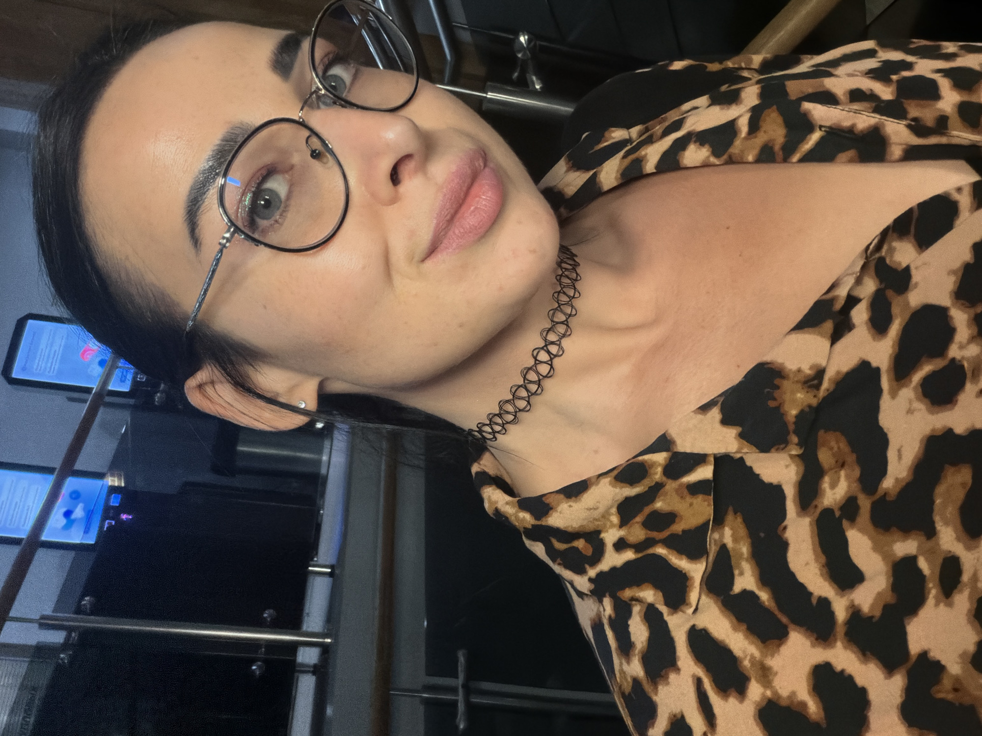 An indoor, waist-up portrait captures a female subject looking directly at the camera with a subtle smile in Saint Petersburg, Russia. The woman, appearing in her 20s or 30s, has dark hair pulled back, revealing a small stud earring in her right earlobe. She wears round-framed glasses with dark and metallic accents. Her eyes are light-colored, and her lips are subtly tinted pink. A black, open-weave choker necklace is visible on her neck. She is dressed in a collared garment featuring a brown, black, and tan leopard print pattern. The background is dark, revealing multiple illuminated rectangular screens displaying blue-toned graphics and text, one of which includes a small purple human figure pictogram. Metallic railings and structural elements are present behind the subject, indicating a modern interior environment.