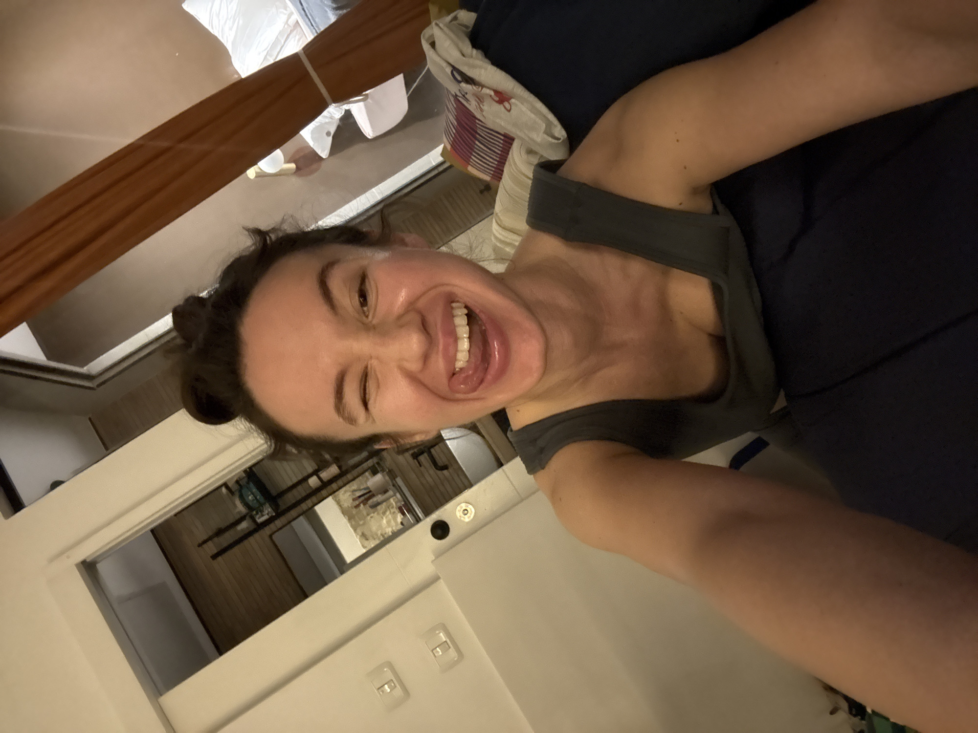 A woman is holding the camera at an angle in what appears to be an apartment interior. She is smiling widely with her tongue visible. She is wearing a gray tank top. The apartment has white walls, a view into another room with wood flooring and what looks like a small kitchen area with appliances visible. The apartment's design seems modern. The exact location in Tel Aviv, Israel is not discernable.