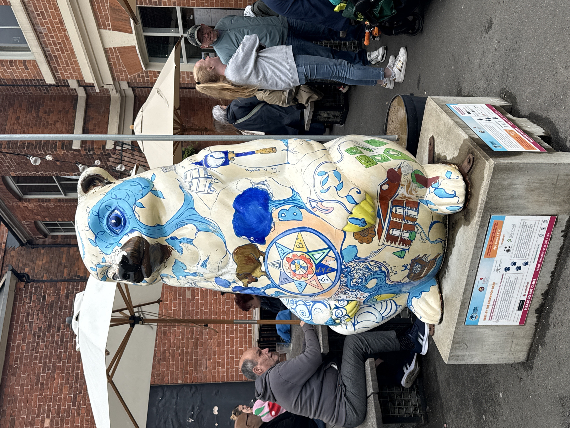 An outdoor scene depicts a large, decorated bear sculpture named "Bentley the Bearwood Bear" positioned on a concrete plinth in what appears to be a public square or courtyard, likely in Bearwood, Sandwell, Black Country, United Kingdom, as indicated by a sign at its base. The sculpture, primarily off-white, is extensively adorned with blue and brown motifs, including a large blue eye and facial markings, a brown bear, and various symbols and sketches such as a bus, a clock tower, houses, and swirling patterns. A sign on the plinth identifies the bear and provides information about "The Big Sleuth" art project, mentioning Sandwell and Positive Activities.
To the left of the bear, a man with a bald head and grey beard, wearing a grey hooded sweatshirt and grey trousers, sits on a wooden bench at a wooden table. He is looking towards his right. A glass is on the table in front of him. Behind him, another person with blonde hair and a pink patterned sweater is partially visible. Further back, a white market umbrella is open over a table.
To the right of the bear, a young woman with long blonde hair in a ponytail stands facing left, conversing with a man to her right. She wears a light grey sweatshirt, blue jeans, and white sneakers. The man wears a dark green jacket and a camouflage baseball cap with "NY" on it, and glasses. He is smiling. Behind them, several other people are present, including a person in a dark coat sitting at a table with another white umbrella in the background.
The background consists of brick buildings with windows, typical of industrial or renovated historical architecture. String lights are visible hanging between a pole and a building on the right. The ground is paved, and a black pot or bin is situated next to the bear's plinth. The overall atmosphere suggests a casual gathering space.