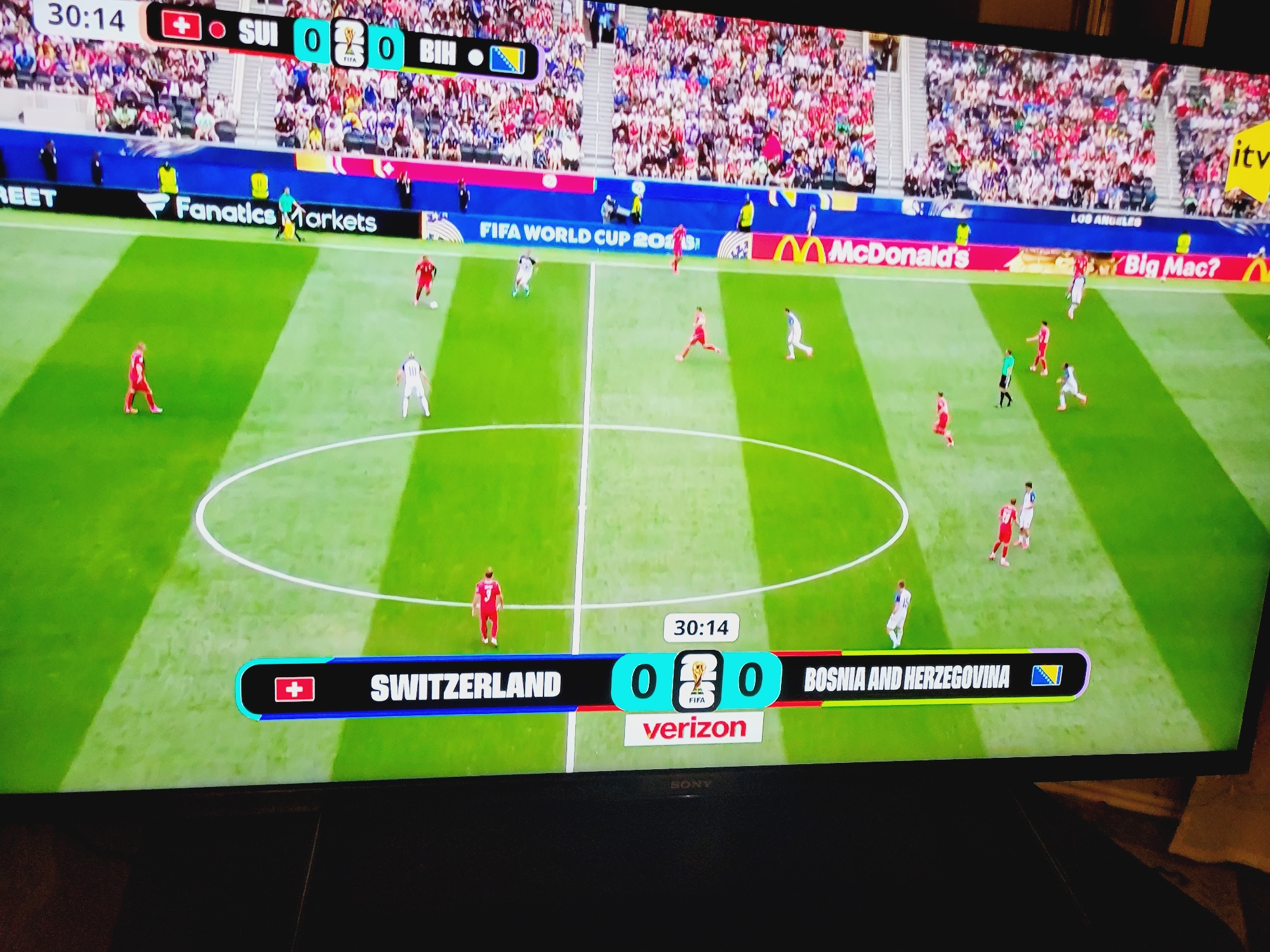 The image shows a soccer match between Switzerland and Bosnia and Herzegovina, as indicated by the score graphic at the bottom of the screen. The score is tied 0-0, and the game is in the 30th minute and 14th second. The match is taking place on a green grass field with white lines marking the boundaries and center circle. Stadium seating filled with spectators is visible in the background. Advertising banners are displayed along the side of the field, including "Fanatics Markets," "McDonald's," and "Big Mac?". A large screen displaying the match details is prominently featured in the foreground. The location is identified as Baku City, Azerbaijan.