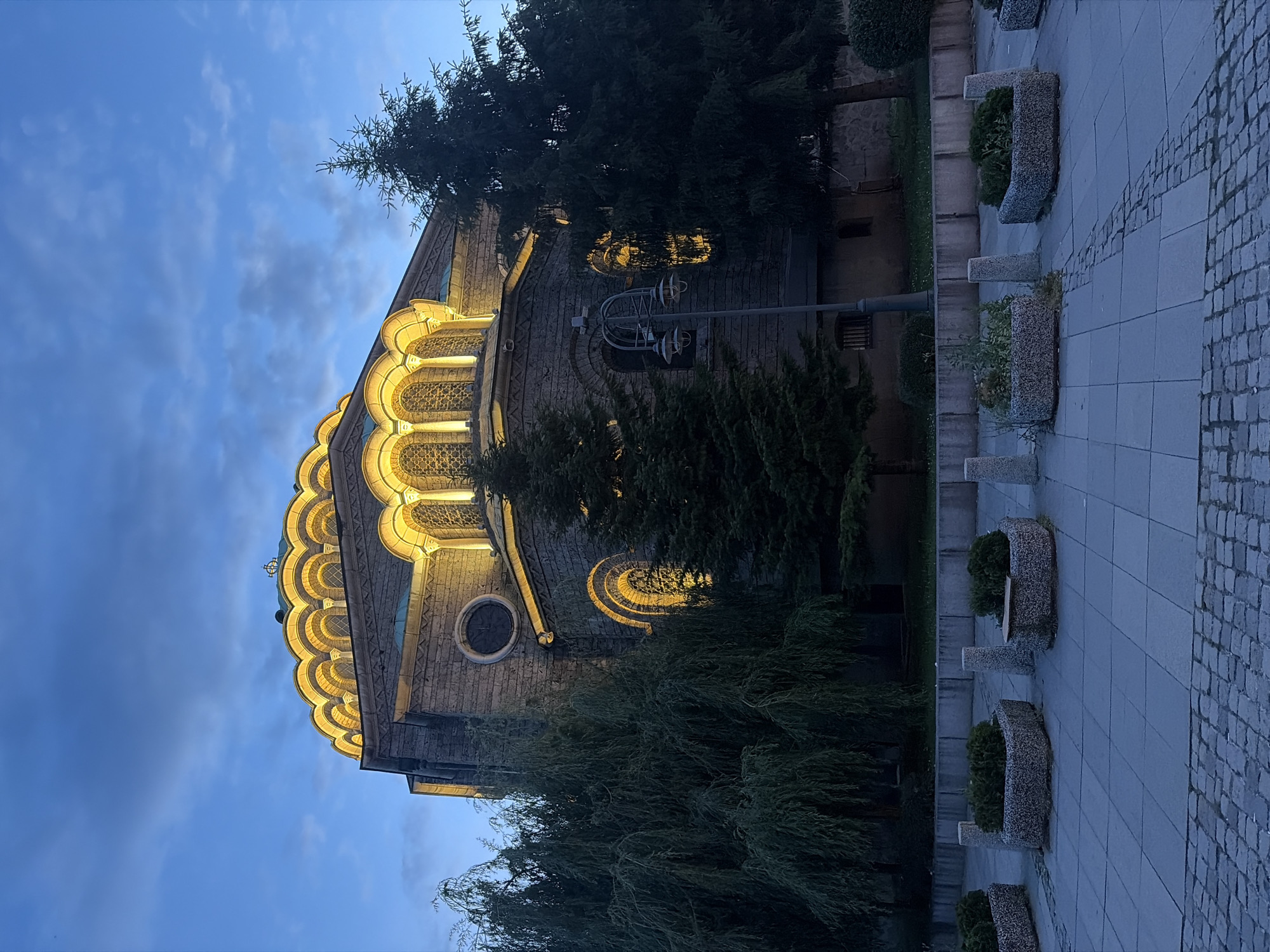 The image shows the Alexander Nevsky Cathedral in Sofia, Bulgaria, at dusk. The cathedral is a large, ornate Orthodox church with a prominent dome and bell tower, constructed from light-colored stone. It is illuminated by warm, golden lights that highlight its architectural details, including arched windows and decorative moldings. The sky above is a deep blue with scattered clouds.
In the foreground, there is a paved plaza with several planters filled with greenery. The planters are made of concrete and have a textured surface. Between the planters, there are cylindrical concrete bollards. To the left of the scene, there are several trees, including a large weeping willow. To the right, there are tall evergreen trees. A street lamp with multiple bulbs stands near the center of the image. The overall scene conveys a sense of grandeur and solemnity, with the illuminated cathedral serving as the focal point. There are no people visible in the image.
