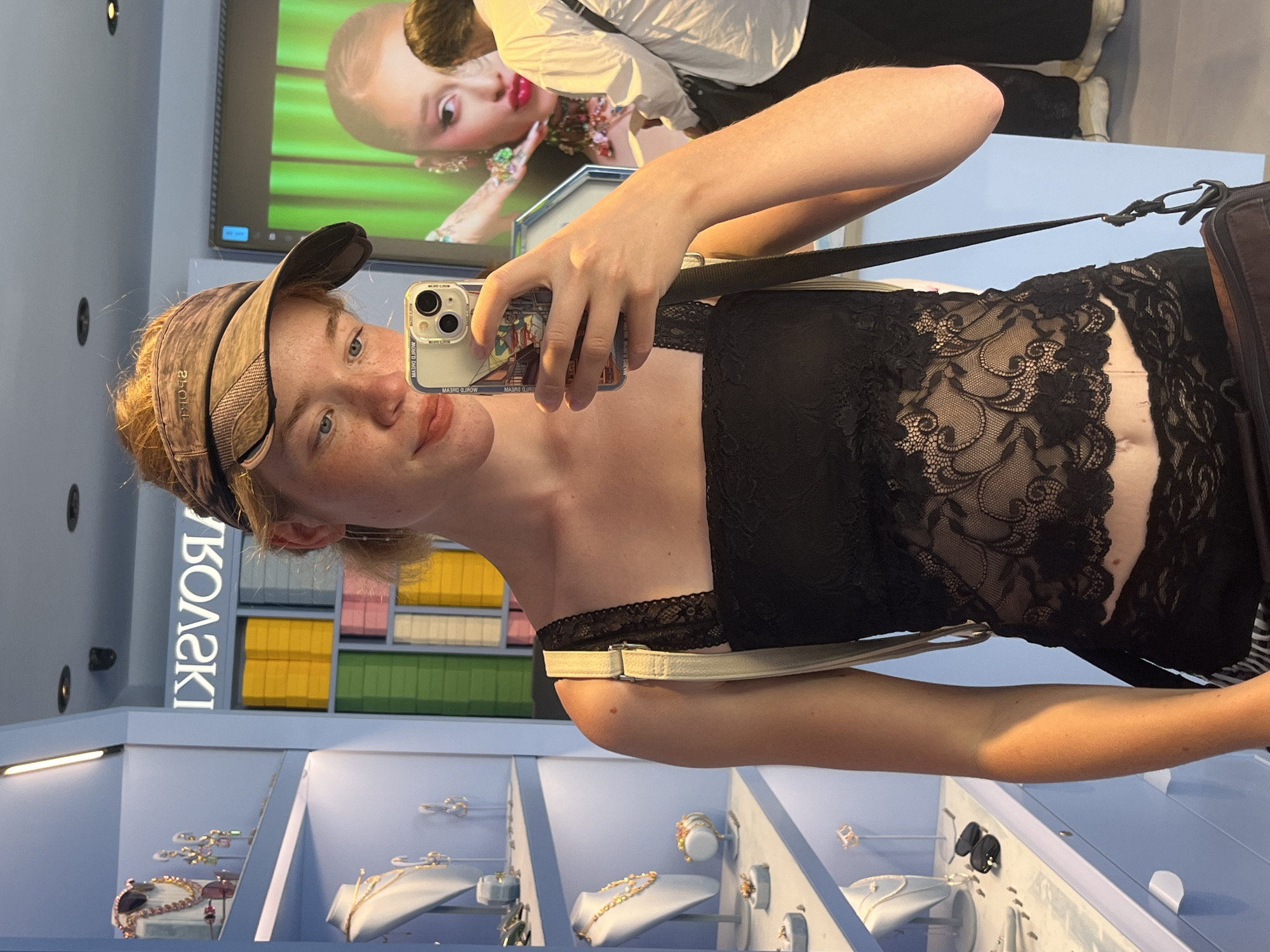 The image captures a young woman taking a selfie in a brightly lit store, likely a jewelry shop, given the displays of necklaces and earrings. She is wearing a black lace camisole, a patterned visor, and her hair is pulled back. The store's interior features pale blue shelving units filled with merchandise and displays. Behind her, a large screen shows an image of a woman with dramatic makeup and jewelry. Another person, wearing a white shirt and dark pants, is visible in the background, partially obscured. The store appears to be located in Budva, Montenegro, suggested by the "SWAROVSKI" lettering visible on a blue wall, a common luxury brand with retail locations worldwide, including in popular tourist destinations like Budva. The overall impression is a candid snapshot within a commercial environment.