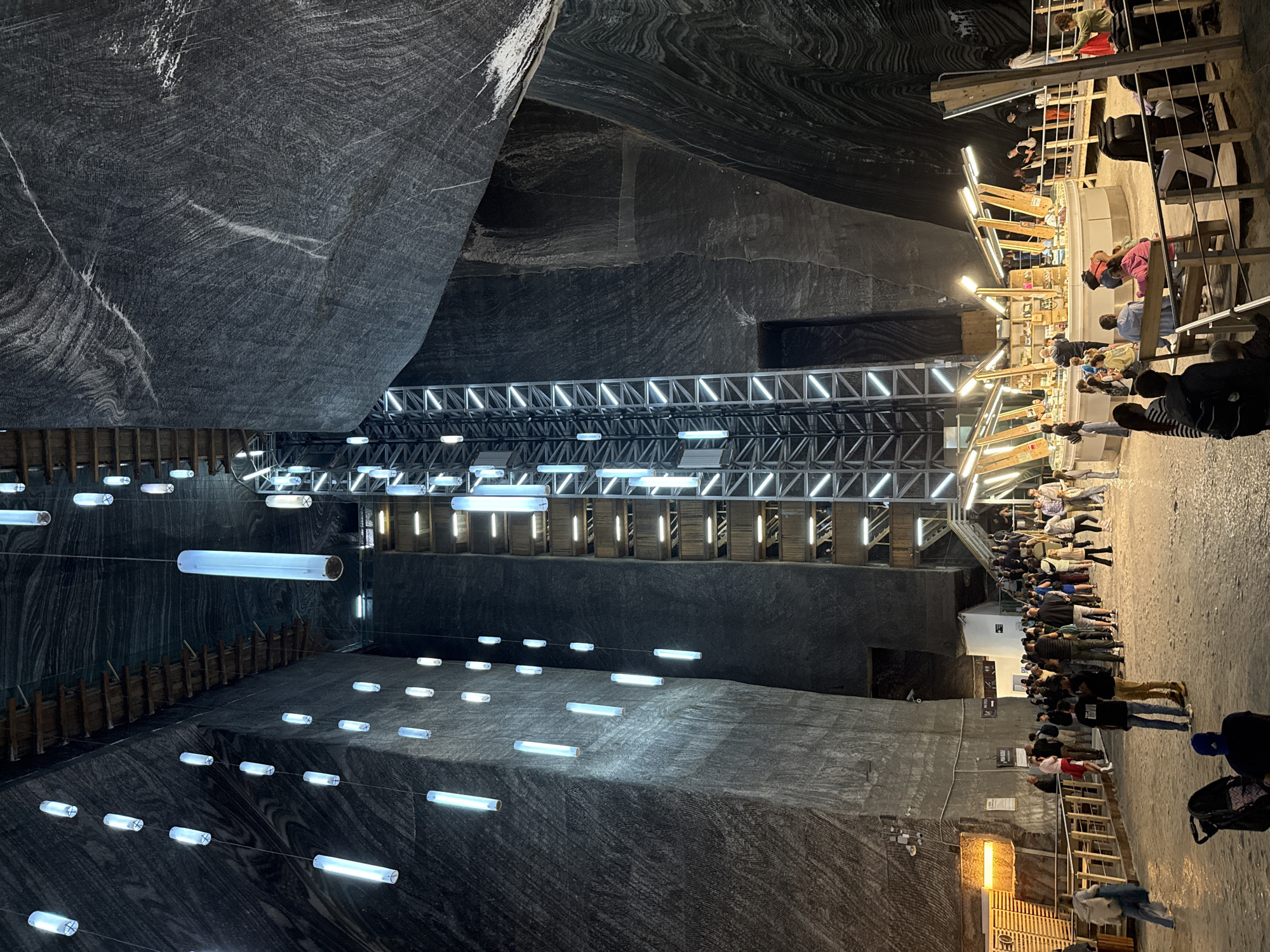 The image depicts a grand, cavernous interior of a salt mine in Turda, Romania. The walls are composed of layered gray salt, with visible striations and some lighter mineral deposits. A central, tall, metallic elevator shaft with multiple levels and illuminated with vertical white lights is the focal point. The ceiling is high and irregular, also made of salt, and is adorned with numerous hanging cylindrical white lights that create an ethereal glow. Wooden scaffolding or structures are visible along the sides and above.
In the foreground, a crowd of people is gathered, appearing to be tourists. They are dressed in casual attire and are walking, standing in groups, and observing the surroundings. Some are near a circular display area that appears to be a shop or exhibition, with products arranged on shelves. A railing separates the walking area from the exhibition space. In the lower foreground, a child's stroller is parked.
The overall impression is one of a vast, illuminated underground space that has been adapted for public access, likely a tourist attraction. The lighting creates a dramatic and somewhat surreal atmosphere within the natural salt formations. The location is identified as the Turda Salt Mine, which is a significant tourist destination in the city of Turda, Cluj County, Romania, not Crairât.