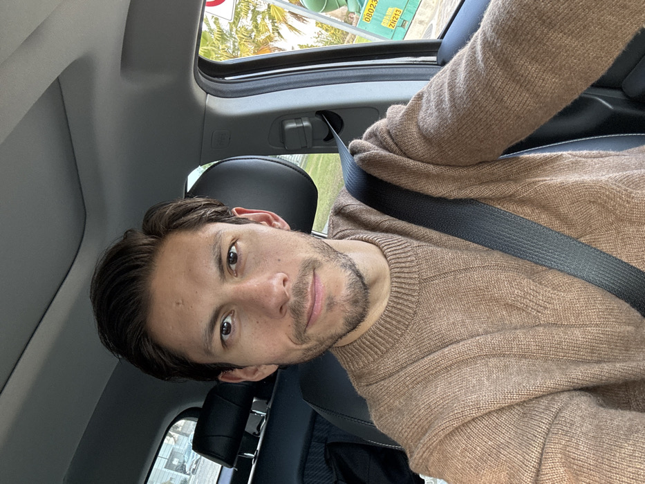 A man with brown hair and a trimmed beard is in a car, wearing a brown sweater and a seatbelt. He's looking at the camera with a slight smile. The car has a sunroof, partially open, revealing greenery and signage. The interior is black. The lighting suggests daytime.