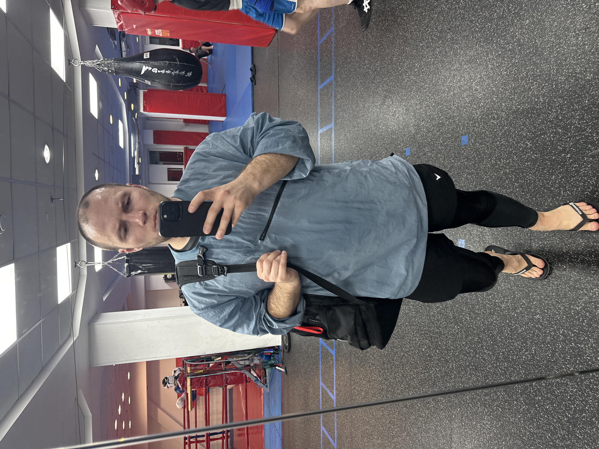 A man is taking a selfie in a gym. He is wearing a light blue shirt, black leggings, and flip-flops. He has a black bag slung over his shoulder. The gym has a boxing ring, punching bags, and blue mats on the floor. The location could be in the city of Krasnoyarsk, Russia, although the specific gym and the city are not definitively discernible from the image alone.