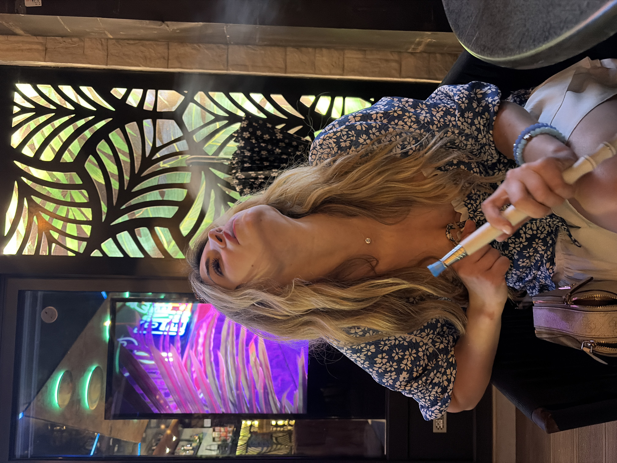 A fair-skinned female with blonde hair is holding a blue-tipped object and looking upward. She is wearing a blue and white floral top, a white top, and black bottoms. She also has a light blue bracelet on her right wrist and a necklace with a pendant. A purse is next to her on the right. Behind her is a decorative panel with cutout leaves. There is a television screen behind the panel showing pink and purple images. The location may be in the city of Budva, Montenegro.