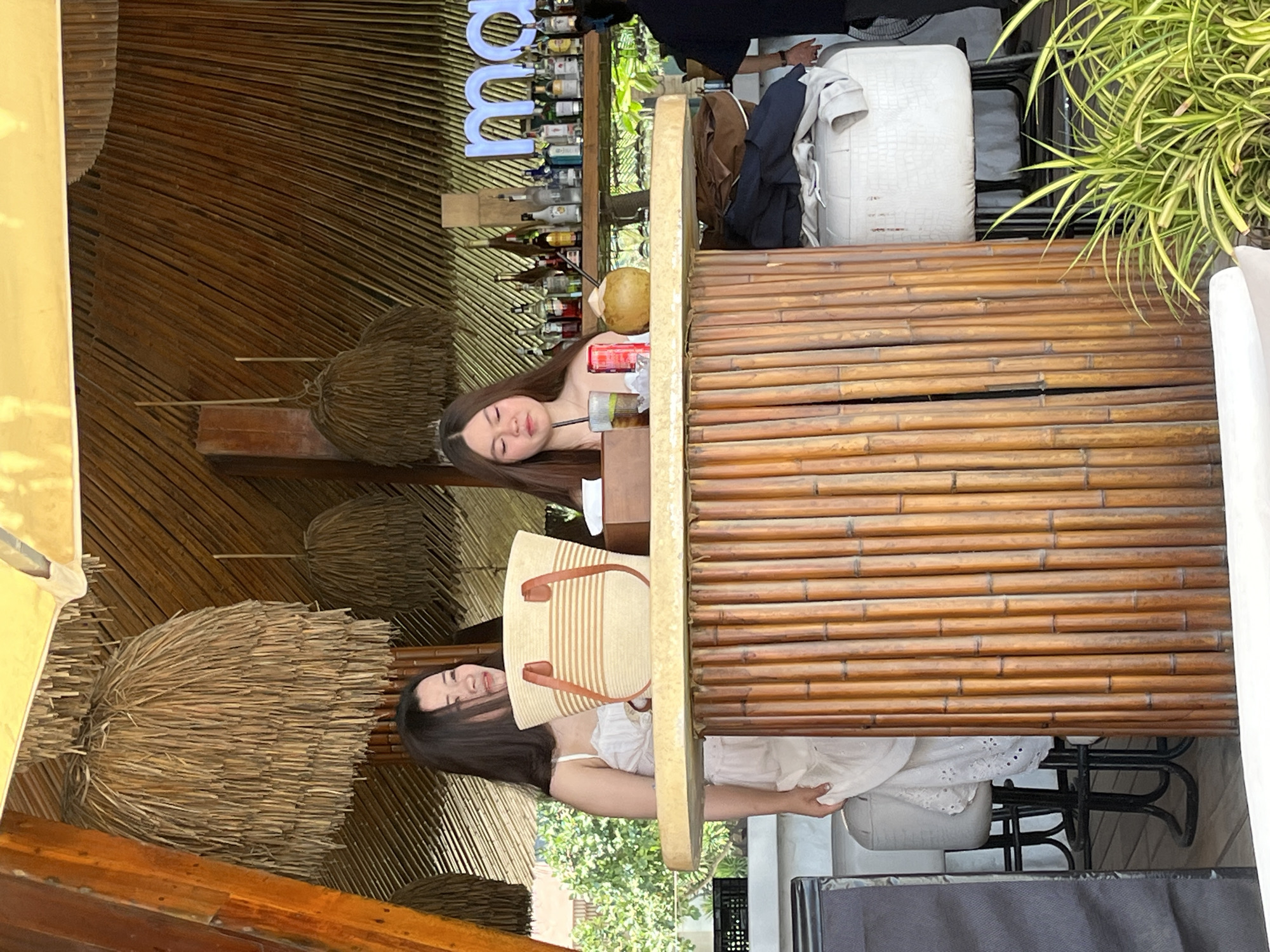 Two women are sitting at a bamboo-covered bar in Đà Nẵng, Vietnam. One woman is resting her head on the bar next to a partially consumed beverage and a coconut. The other woman is holding a striped tote bag. The bar is stocked with bottles of alcohol. The environment includes straw umbrellas, tropical foliage, and seating.