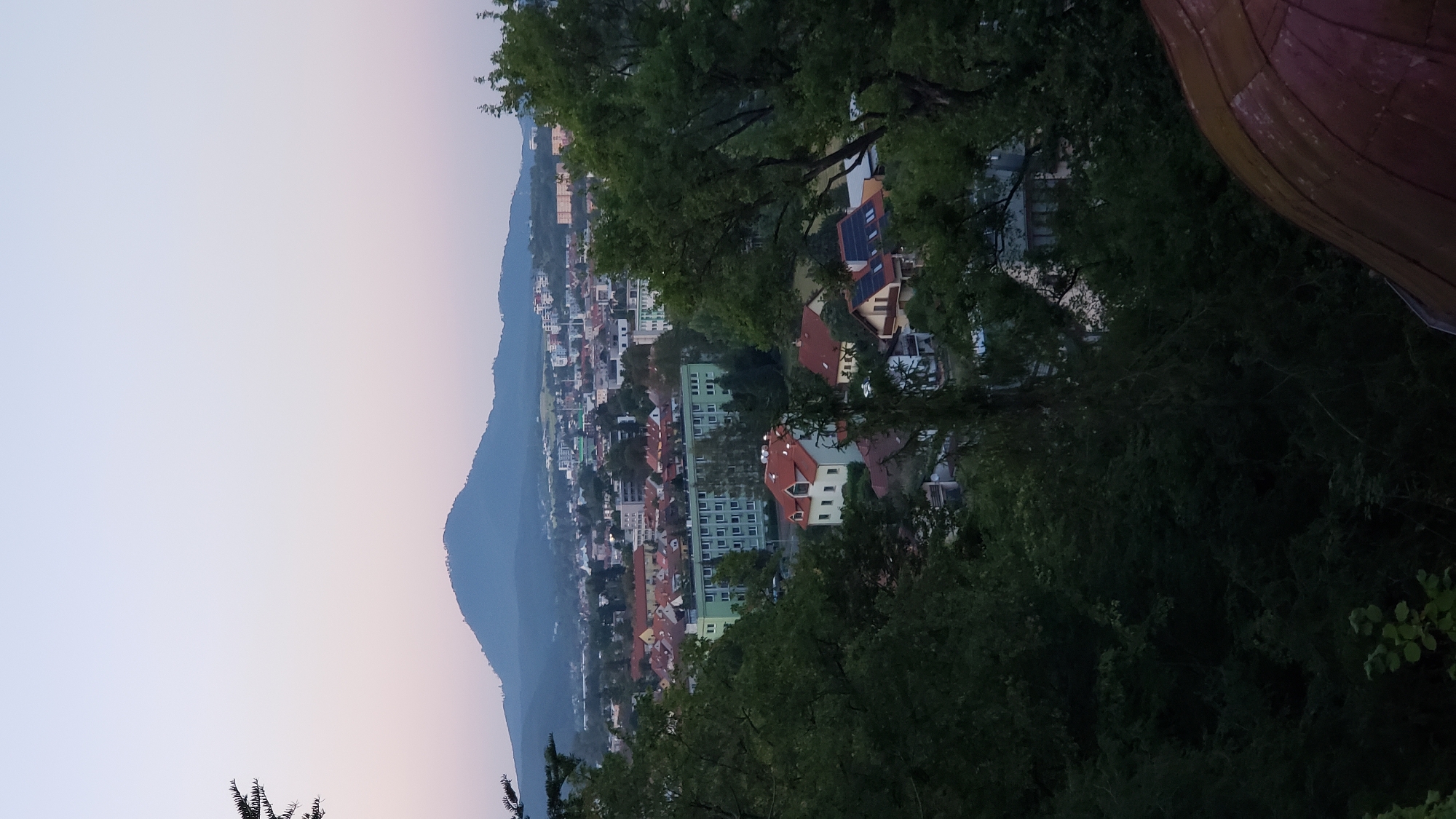 The image shows a panoramic view of the city of Prešov, Slovakia, at dusk. In the foreground, dense green foliage obscures much of the view, with a glimpse of a reddish-brown domed structure at the bottom right. Beyond the trees, the city unfolds with a mix of residential buildings, some with red-tiled roofs, and larger apartment blocks, notable for their green facade. Further in the distance, a prominent hill or mountain dominates the skyline, its slopes covered in dark green trees and its summit appearing to have some structures. The sky above is a gradient of pale pink and blue, characteristic of twilight. The overall impression is a tranquil cityscape viewed from an elevated position overlooking the residential areas and a natural landmark.