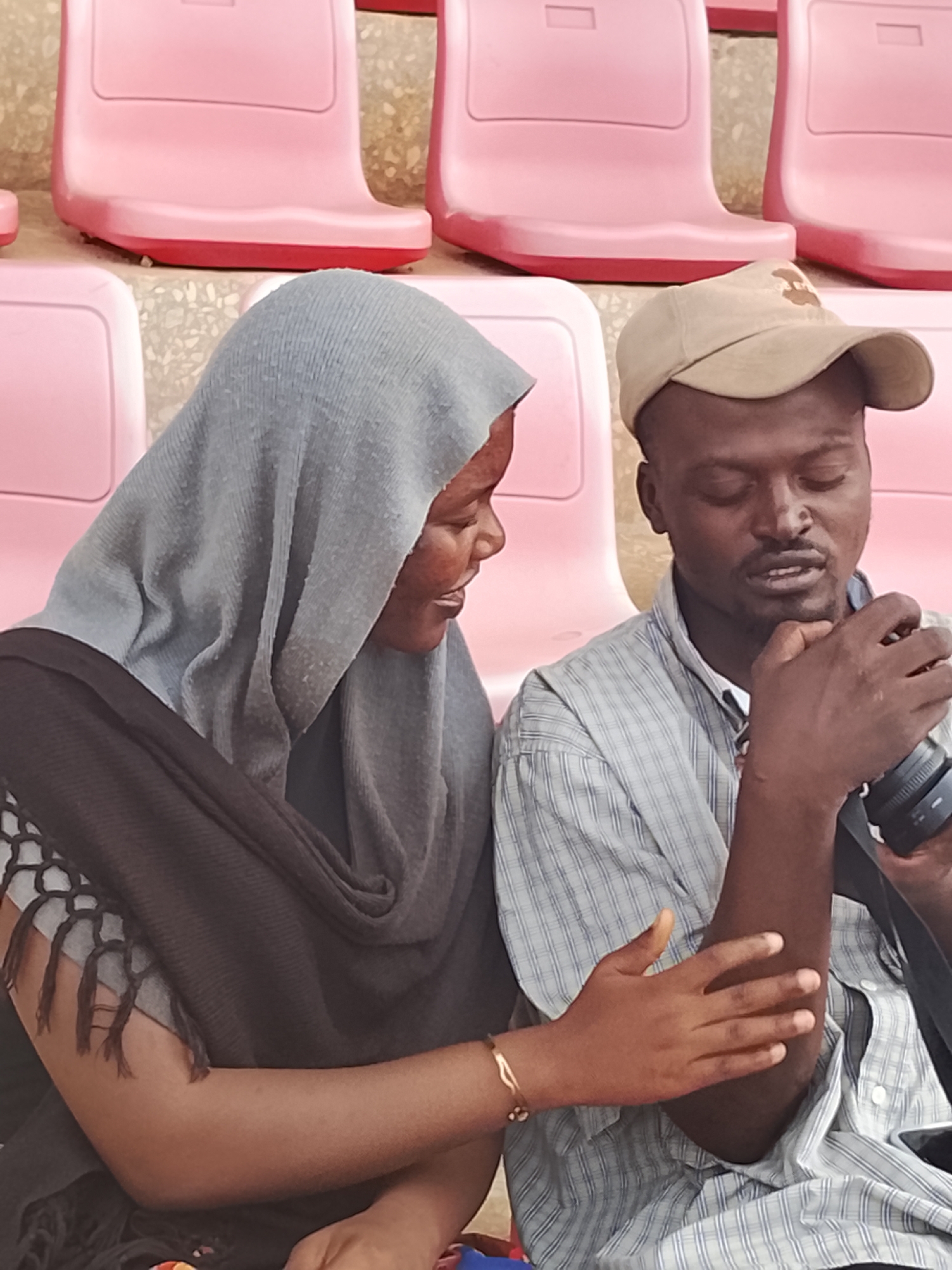 In a medium close-up shot, two individuals are seated, likely in a stadium or arena, indicated by the rows of empty pink plastic seats in the background. The scene takes place in Zawan, Nigeria.
On the left is a woman, seen in profile, wearing a light grey hijab and a dark shawl over a lighter, possibly patterned top. Her face is turned towards the man, and she appears to be smiling subtly, her expression engaged. She wears a delicate gold-colored bracelet on her right wrist, and her right hand is resting gently on the man's left forearm.
On the right is a man, wearing a light-colored, possibly blue and white, plaid button-up shirt and a beige baseball cap. He is holding a black camera with a lens in his hands, and his gaze is directed downwards towards the camera, suggesting he is operating it, reviewing footage, or explaining something about it. His mouth is slightly open as if speaking or concentrating.
The interaction between the two suggests a shared moment of interest or instruction related to the camera. The lighting indicates it is daytime. No discernible text is visible in the image.