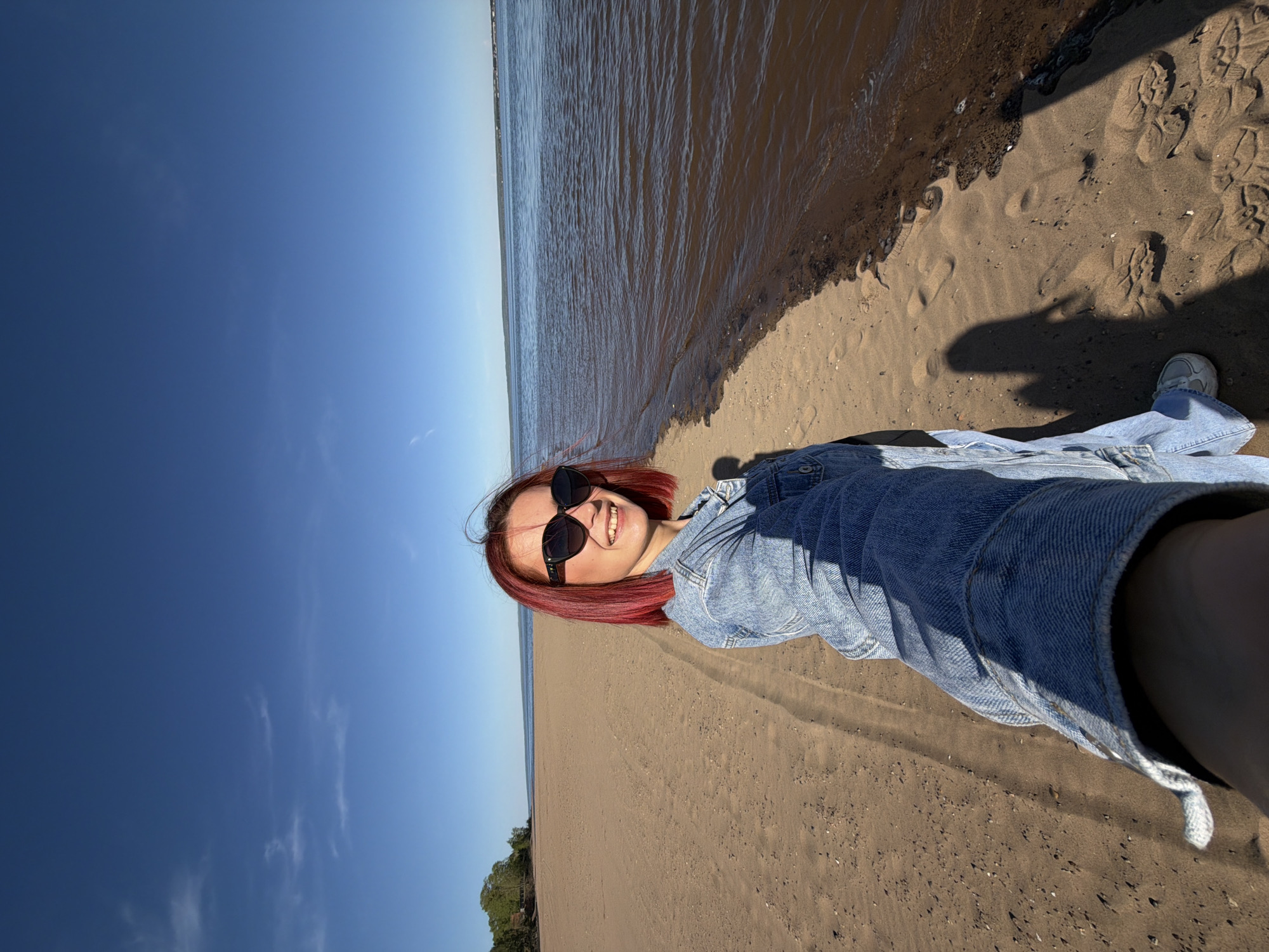 A young woman with auburn hair, wearing sunglasses and a denim jacket, is standing on a sandy beach. She is likely taking a selfie. The water is to the right, and a clear blue sky with some clouds is on the left. The beach appears to be located on a river, rather than an ocean. A small line of trees is on the distant shore. Based on the photo metadata, the location is Noviy, Russia.