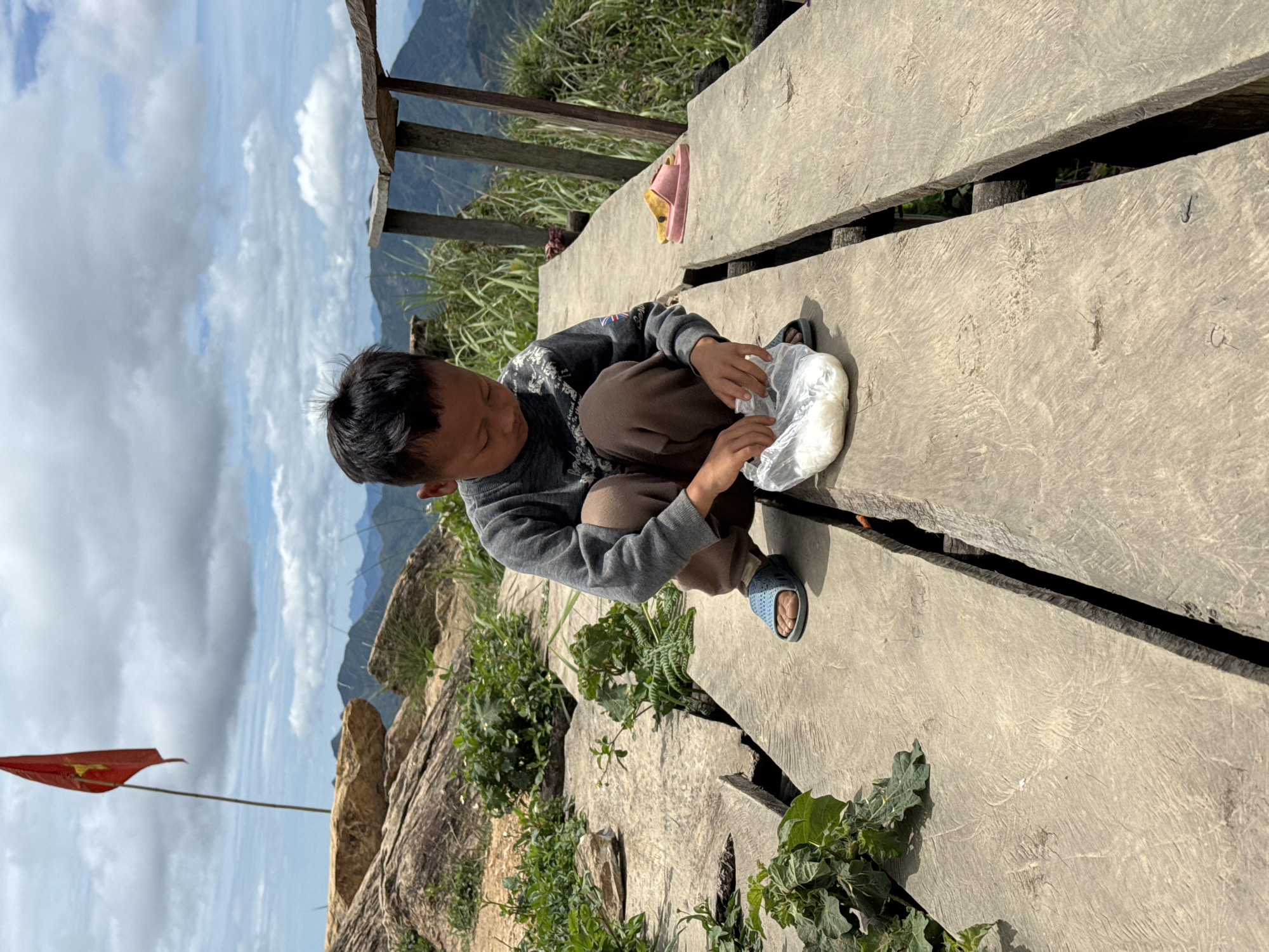 A young boy is crouched on a wooden pathway on a mountaintop in Ta Xua, Vietnam. He is wearing a grey long-sleeved shirt and brown pants, and blue sandals. He is looking down at a clear plastic bag filled with white substance, possibly food, in his hands. A Vietnamese flag is planted on a rocky outcrop behind him, and a wooden railing is visible to his right. The background consists of rolling green hills and a cloudy sky. The wooden pathway is weathered and cracked, with vegetation growing around it. A pair of pink and yellow flip-flops are placed on the pathway to the boy's right.