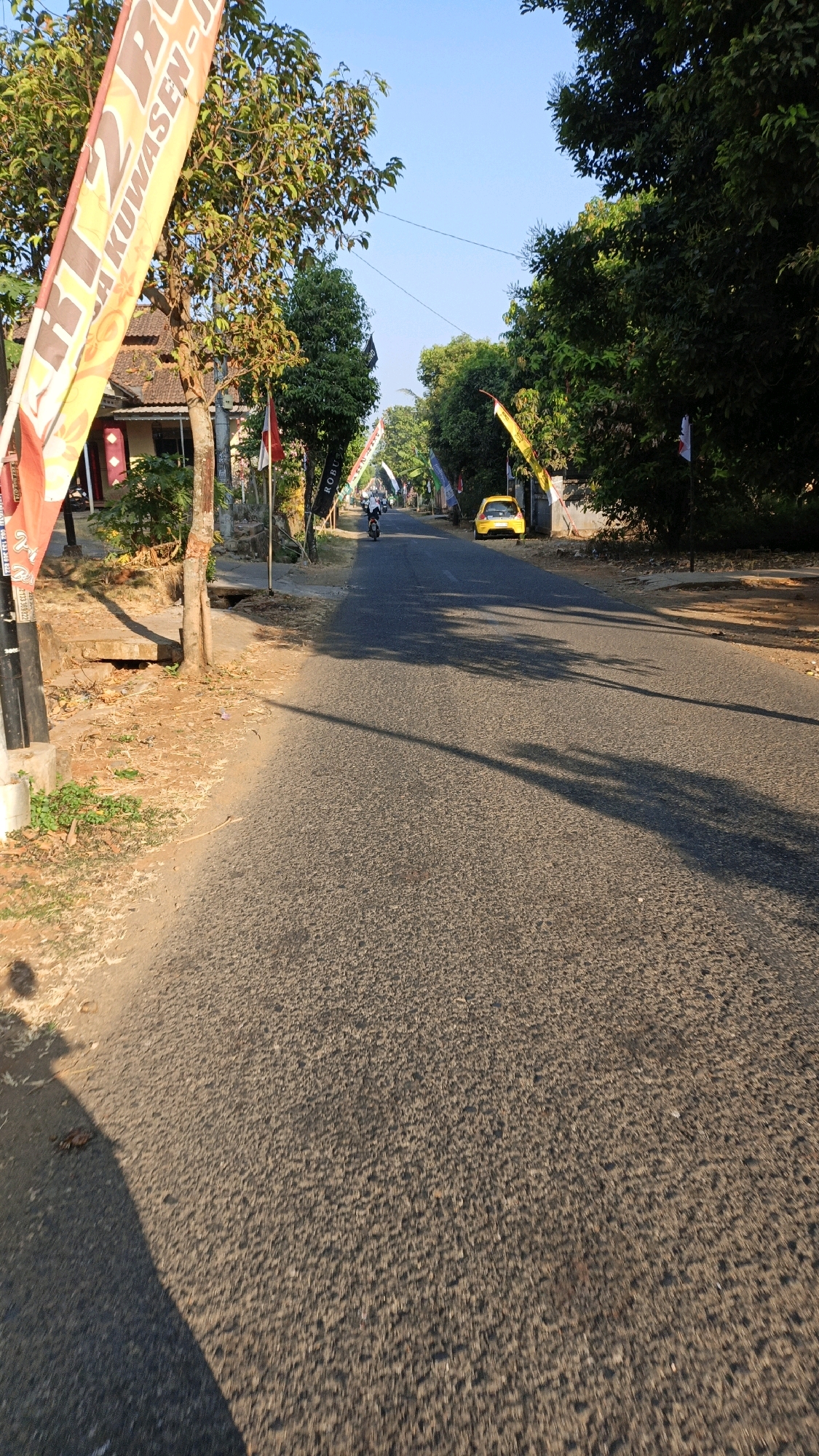 The image depicts a road in the city of Kuwasen, Indonesia, under a clear blue sky. The road is paved and appears to be a two-lane street lined with trees and vegetation. Several flags are visible along the side of the road, some with text on them. In the foreground on the left, a shadow is cast from an unseen object. Further down the road, a yellow car is parked on the right side. A person is seen riding a motorcycle in the distance, heading away from the camera. Buildings are visible on the left side of the road behind some trees and flags. The scene suggests a quiet street in a residential or semi-rural area.