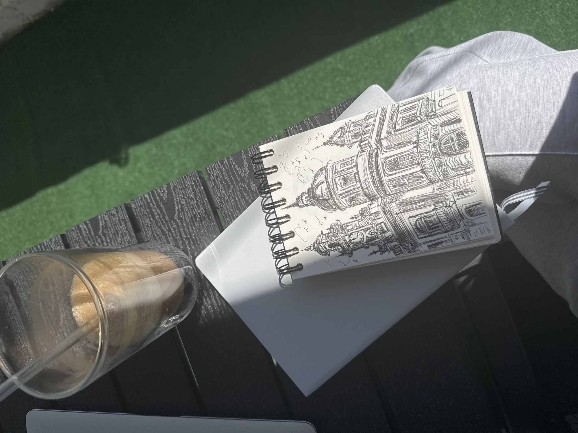 The image captures a high-angle, medium shot of a notebook resting on top of a white laptop, with a glass containing a beverage and a straw visible to the left. The background appears to be an outdoor setting with an artificial grass surface. The notebook contains a detailed pen and ink drawing of an architectural structure resembling a building with domes and clock towers. Parts of the drawing suggest it is Alexander Nevsky Cathedral, in Sofia, Bulgaria. A light-colored hooded garment is draped slightly behind the laptop and notebook. The composition is bathed in natural daylight, creating shadows that suggest an outdoor setting on a sunny day. The shot is composed in a way that the elements of drawing, drink, technology, and casual clothing coexist, suggesting a moment of creative activity outdoors.