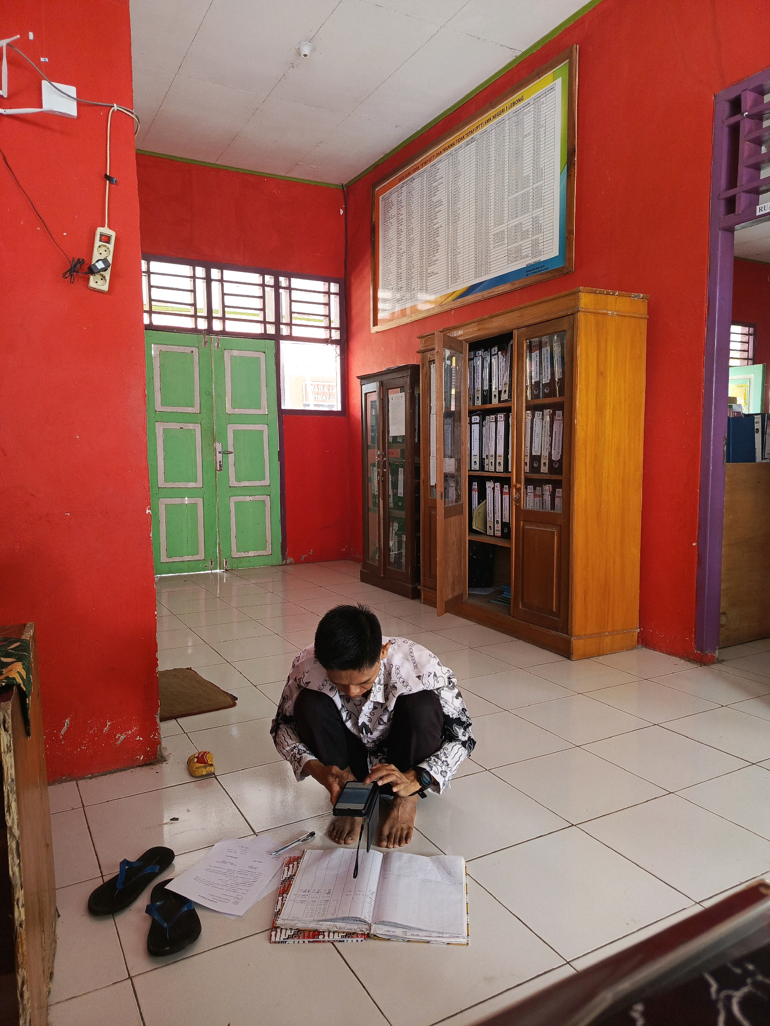 The image depicts an indoor scene, likely a school or office environment, with bright red walls and a tiled floor. A man, wearing a patterned white shirt and dark pants, is crouching on the floor, engaged with his mobile phone. He appears to be in the process of looking at or using the device, with one hand holding it and the other resting near it.
Scattered around him on the floor are several items, including a pair of black flip-flops, a document with a pen, and an open book, suggesting he may be studying or working. A small yellow figurine is also visible near his feet.
In the background, a wooden cabinet filled with folders and binders stands against the red wall. Another similar cabinet is partially visible to the left. A large framed chart or notice is mounted high on the wall. Green double doors with white trim are also present, suggesting an exit or entrance. The overall setting appears to be a functional space, possibly a classroom or administrative area within a building in Tik Jeniak, Indonesia.