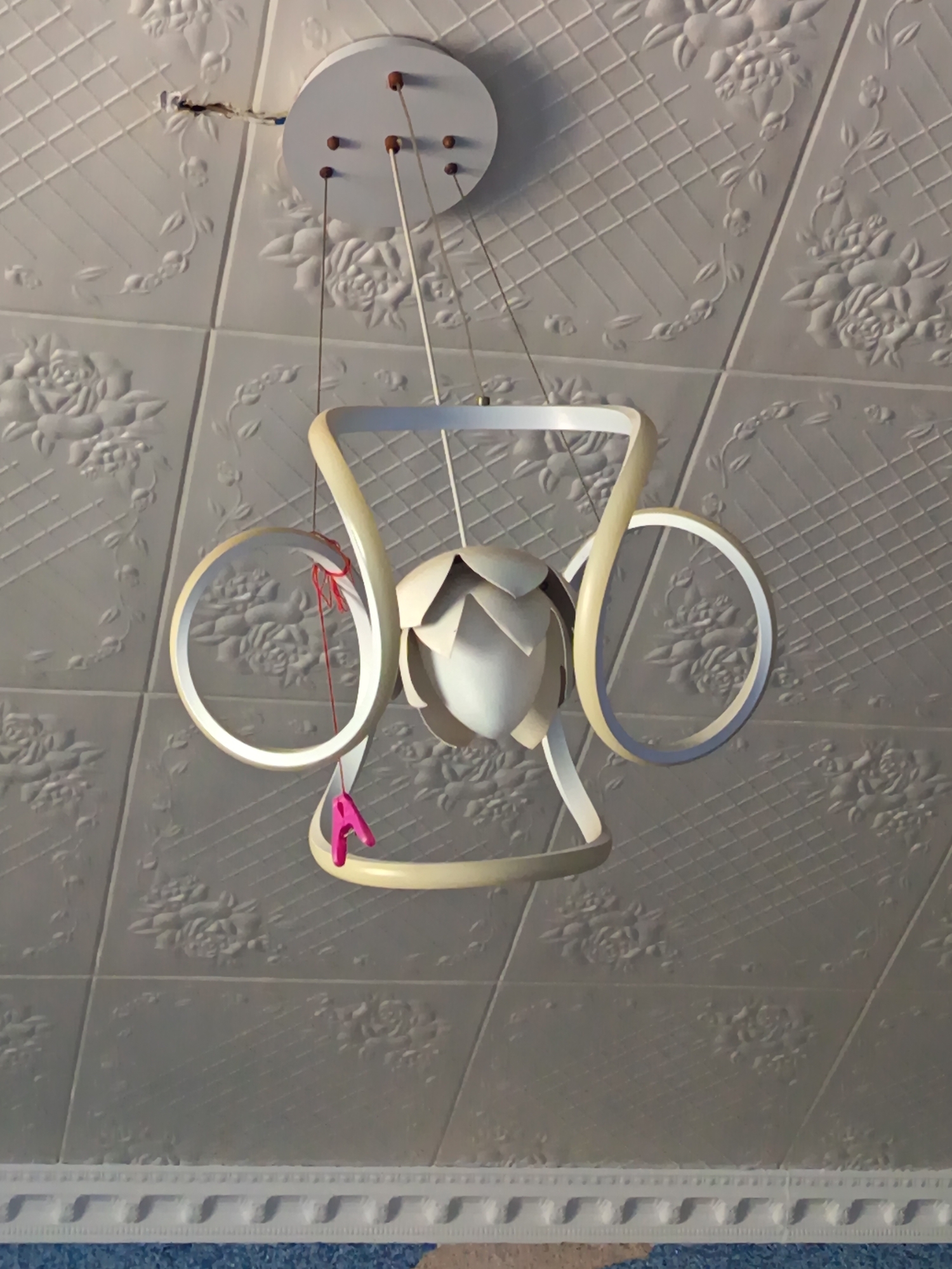 A modern chandelier with a cream-colored frame hangs from a white, textured ceiling, which is patterned with embossed floral designs. The chandelier features two large, curved arms that form rings, and a central bulbous diffuser that resembles a flower bud. A thin red string is attached to one of the arms, and a bright pink clothespin is clipped onto another. The ceiling is a grid of square panels, each with a symmetrical rose pattern and cross-hatching. At the top left of the image, exposed electrical wires are visible, indicating a recent installation or repair. The lower portion of the image shows the edge of the ceiling with decorative molding, below which is a glimpse of a blue textured surface. The location is the city of Shirin, Uzbekistan.