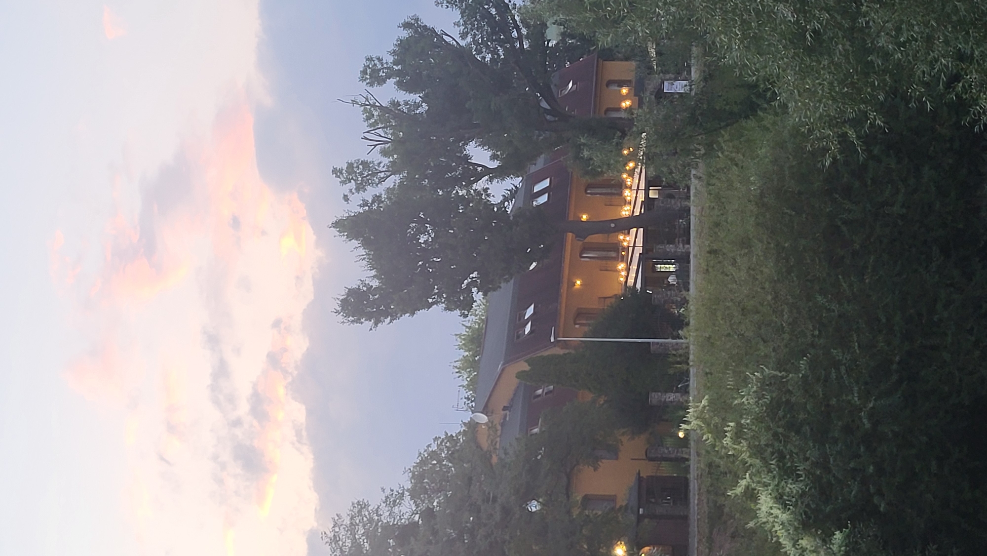 The image shows a building with a yellow facade and a dark red roof, likely a hotel or restaurant, situated in Prešov, Slovakia. The building features multiple windows, some with skylights, and an outdoor seating area illuminated by string lights. Large trees flank the structure, and lush greenery is visible in the foreground. The sky above is filled with soft, pastel-colored clouds, suggesting it is either dawn or dusk. The scene conveys a tranquil and inviting atmosphere.