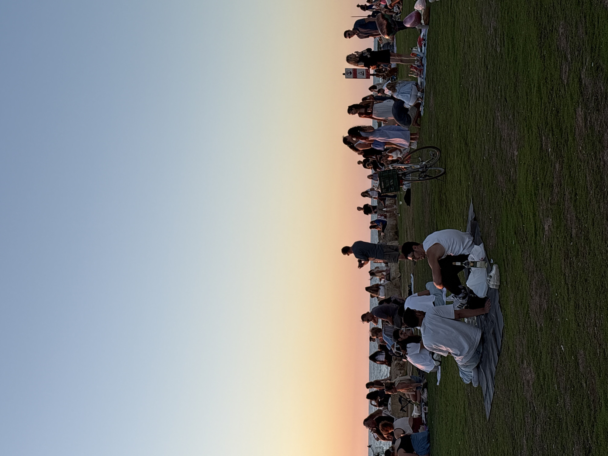 The image shows a group of people picnicking on a grassy area near the sea in Tel Aviv, Israel, during sunset. The sky transitions from a clear blue at the top to a warm orange and pink hue near the horizon. In the foreground, two men are sitting on a picnic blanket, engaged in conversation. One man is wearing a white t-shirt and light-colored pants, while the other is wearing a white tank top and dark pants. A bottle of wine and a pair of sneakers are visible on the blanket. To the left, a bicycle with a basket is parked on the grass.
Further back, a diverse group of people are scattered across the scene, many sitting on blankets or rocks. Some are interacting with each other, while others are looking out towards the sea or at their phones. Several individuals are standing, including a man taking a photo with his phone and another man standing near a signpost. The background features the ocean and a rocky shoreline, with more people gathered along the water's edge. The overall atmosphere appears relaxed and social, with people enjoying the evening outdoors.