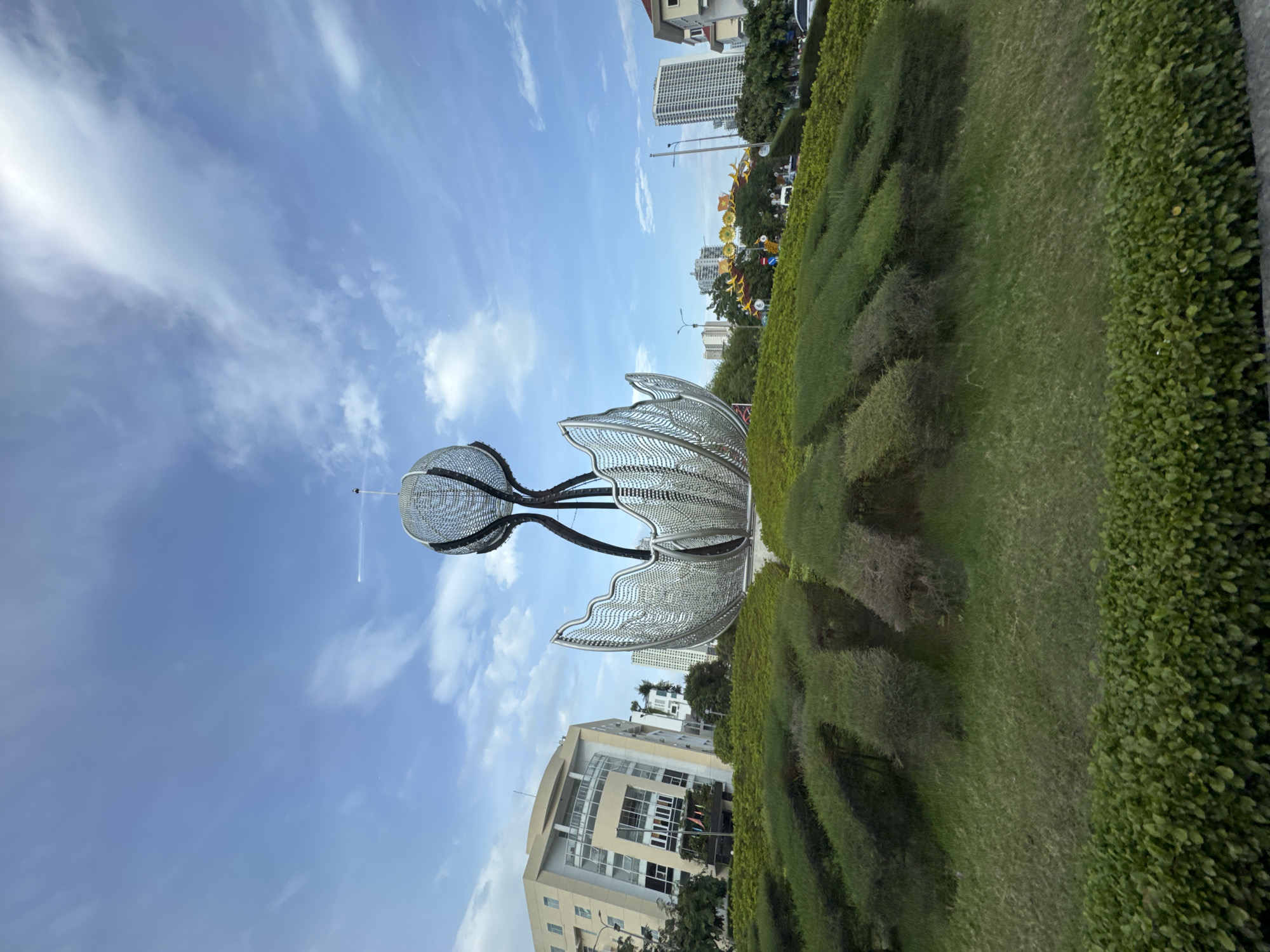 The image depicts a large, modern metal sculpture resembling a lotus flower, situated on a landscaped roundabout in Nha Trang, Vietnam. The sculpture, composed of intricate metalwork, stands tall against a backdrop of a bright blue sky with scattered white clouds. In the background, several tall, modern buildings and some smaller residential structures are visible, indicating an urban environment. The roundabout itself is covered in lush green grass and meticulously trimmed bushes, arranged in a pattern that enhances the visual appeal of the sculpture. The overall scene suggests a well-maintained public space, likely a prominent landmark within the city.