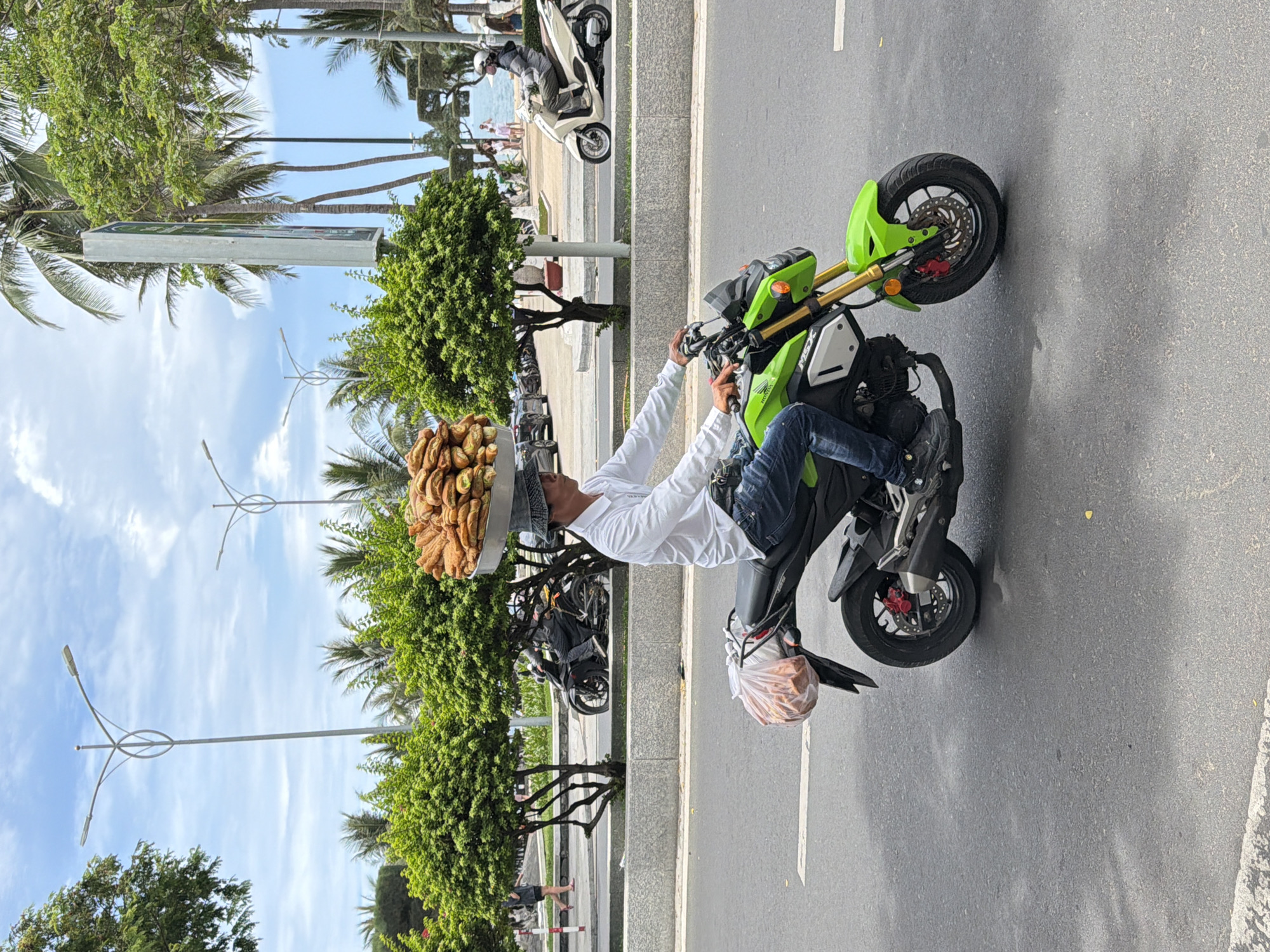 A street scene in Nha Trang Ward, Vietnam depicts a man precariously balancing a large tray of fried pastries on his head while riding a green and black motorcycle. The man, wearing a white shirt and jeans, is driving on a paved road alongside a sidewalk lined with trees and lampposts. A second motorcycle is visible in the background, parked on the sidewalk, with additional people in the distance near the beach. A plastic bag appears to be hanging from the motorcycle's rear. The sky is partly cloudy.