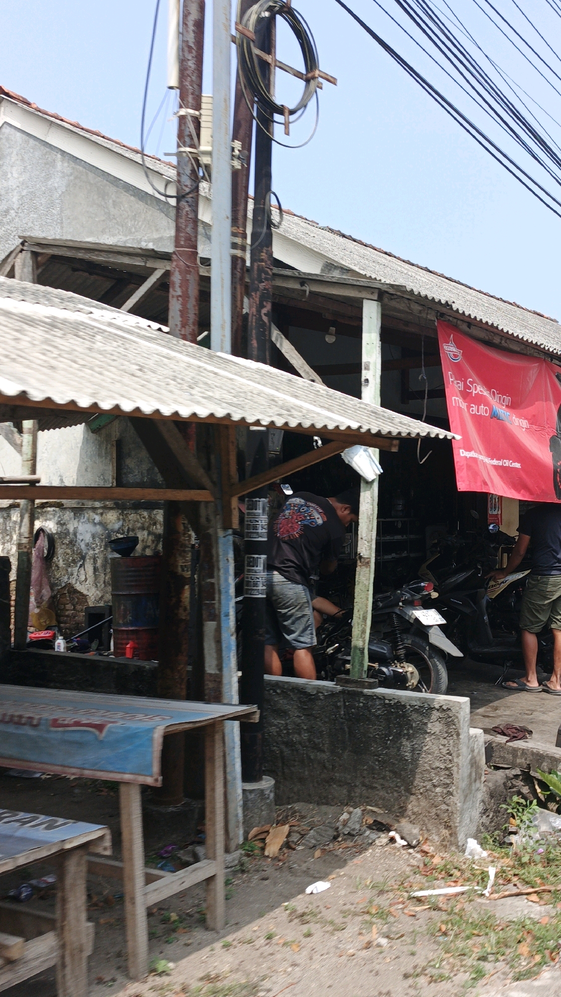 The image depicts an outdoor repair shop in Desa Wonoharjo, Indonesia. In the center, a man wearing a black t-shirt with a graphic design on the back and denim shorts is working on a motorcycle. To his right, another person, only their legs and lower torso visible, wearing shorts, stands near a motorcycle. A red banner with Indonesian text is hung above the work area. The shop appears to be a makeshift structure with a corrugated metal roof supported by wooden beams. Various objects are scattered around the site, including barrels, tools, and debris. In the foreground, two wooden tables with blue coverings are positioned. The environment suggests a rural or semi-urban setting with a dirt ground and vegetation. Electrical wires are visible overhead, supported by utility poles. The sky is clear and blue.